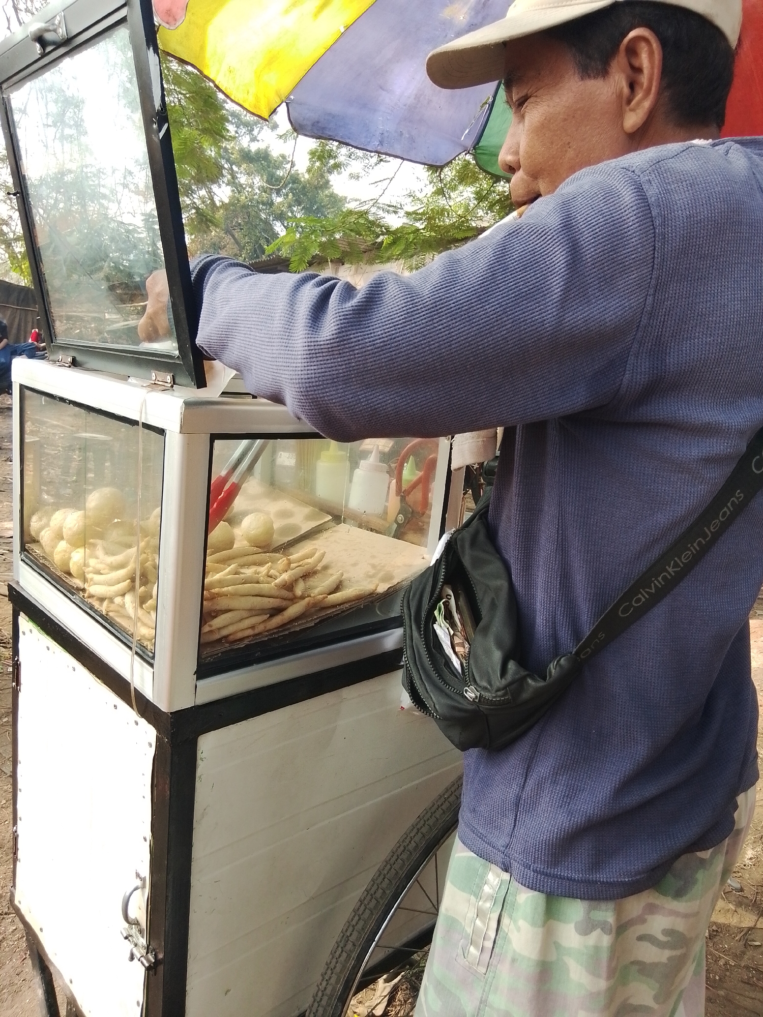 In Bencongan, Indonesia, a person wearing a light-colored cap, a blue long-sleeved shirt, and camouflage pants stands next to a food cart. The cart has a glass display case filled with fried snacks. The person is holding tongs and appears to be attending to the food. A black crossbody bag with the "Calvin Klein Jeans" logo is slung across their chest. The environment suggests an outdoor setting with trees visible in the background and a bicycle wheel on the right side of the frame.