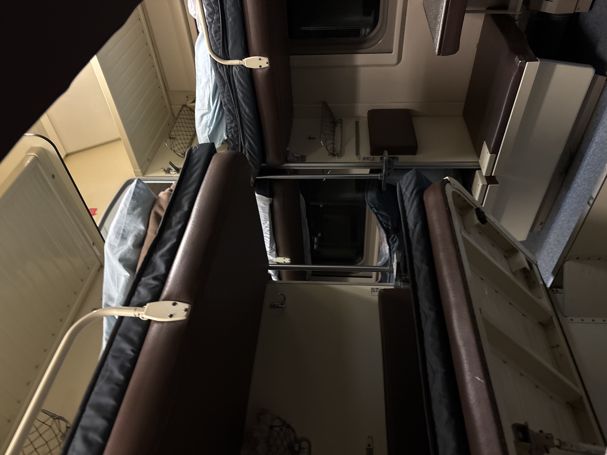 The image shows the interior of a train compartment. The focus is on two sets of seats upholstered in brown, facing each other. There are sleeping bags and bedding stored above the seats. Walls are light-colored. A reading lamp is mounted above each seat. There are windows and shelves visible. The image depicts a typical scene inside a sleeper car on a train. Шемордан, Russia is provided as a location, although there is nothing in the image to independently confirm this.