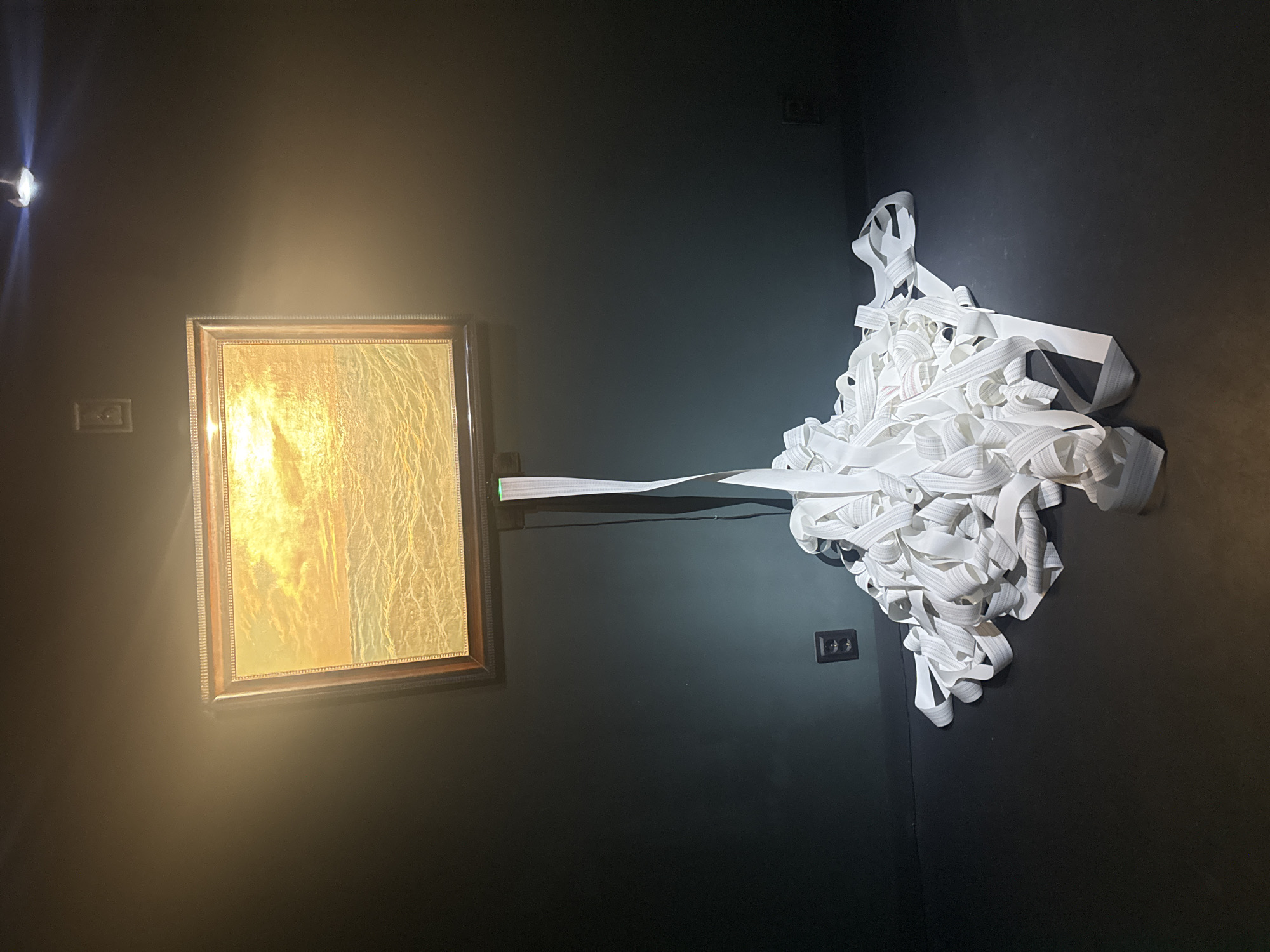 The image shows an art installation in what appears to be a gallery in Sofia, Bulgaria. A framed golden-hued picture hangs on a dark wall. A long white textile strip extends from the top of the frame to a large, chaotic pile of the same material on the right. A spotlight illuminates the picture. Electrical outlets are visible on the wall.
