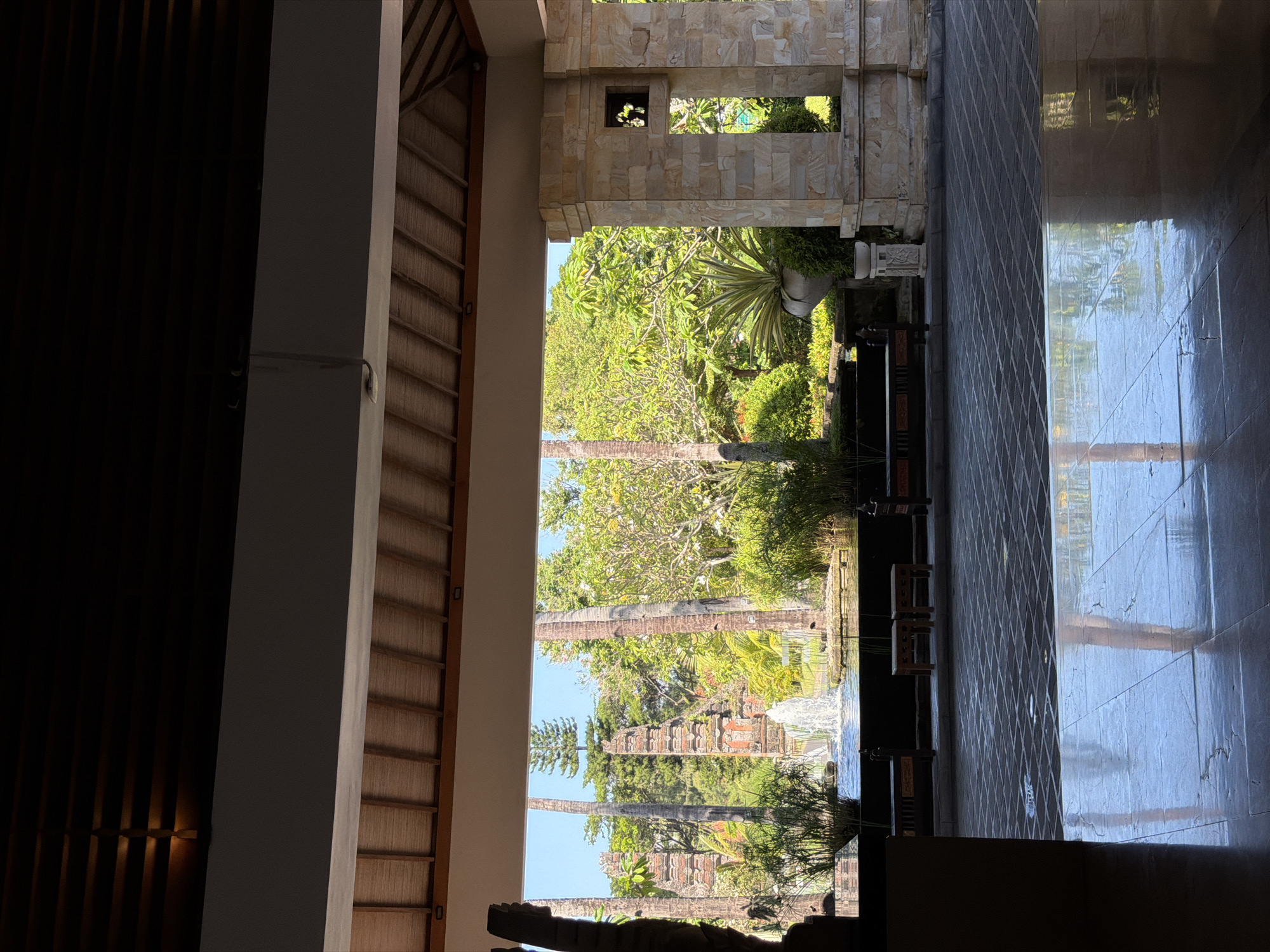 The photograph shows a view from inside a building in Kuta, Indonesia, looking out to a lush garden. The interior is composed of a dark, shadowed area to the left with a textured wall, transitioning to a brighter, sunlit stone archway. Through the archway, a vibrant garden scene unfolds, featuring palm trees, a waterfall, and various tropical plants. The foreground includes dark furniture and decorative elements. The right side of the image displays a reflective tiled floor, mirroring the garden view and suggesting the presence of water or a polished surface. The overall atmosphere is one of serenity and tropical luxury.