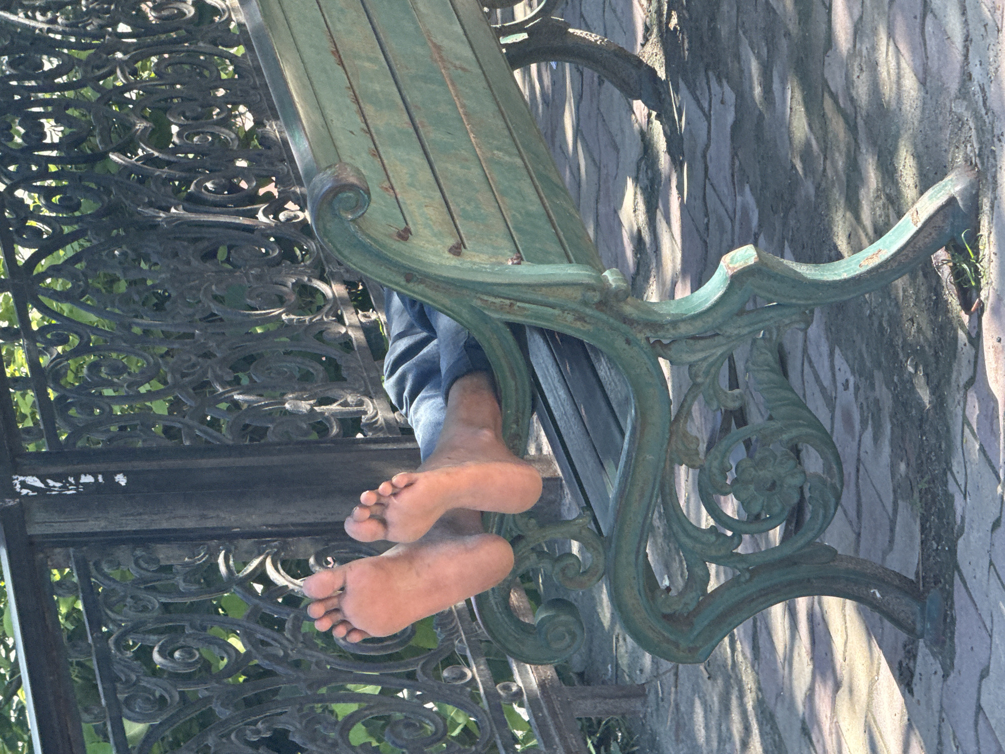 A person's bare feet rest on the back of a weathered, green metal park bench in Rishikesh, India. The bench features ornate scrollwork on its legs and frame. The person is wearing blue jeans. In the background, a dark, decorative metal fence with intricate scrollwork is visible, with green foliage behind it. The ground beneath the bench is paved with textured, light-colored bricks. Sunlight casts dappled shadows on the ground and the bench.