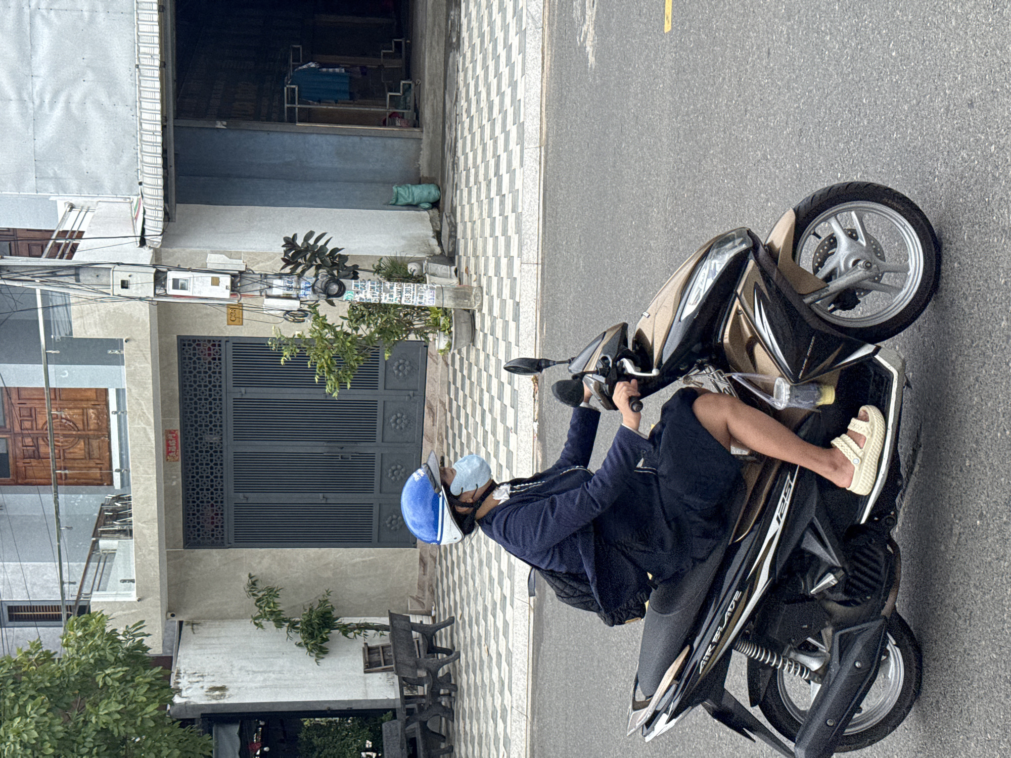 A person wearing a blue helmet and a light blue face mask is riding a dark brown and silver Honda Air Blade 125 scooter on a paved street. The person is wearing a dark blue jacket, dark shorts, and light-colored sandals. A black backpack is visible on the person's back. A plastic cup with a yellow liquid is attached to the scooter. The street is bordered by a checkered tiled sidewalk, a building with a gray facade and a dark gray gate, and a utility pole with numerous wires and electrical boxes. The building appears to be a residential or commercial establishment. The sky is overcast. The scene is likely located in Phan Thiết, Vietnam, given the style of architecture and the prevalence of scooters.