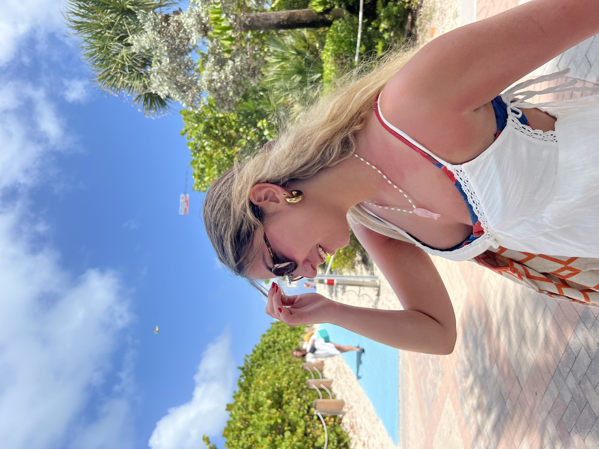 A woman in a white dress stands outdoors. She has blonde hair and wears sunglasses. The scene includes a blue sky with clouds, trees, and a paved walkway. In the background, a person in a white dress is on a blue surface with the beach. This may be located in Miami Beach.