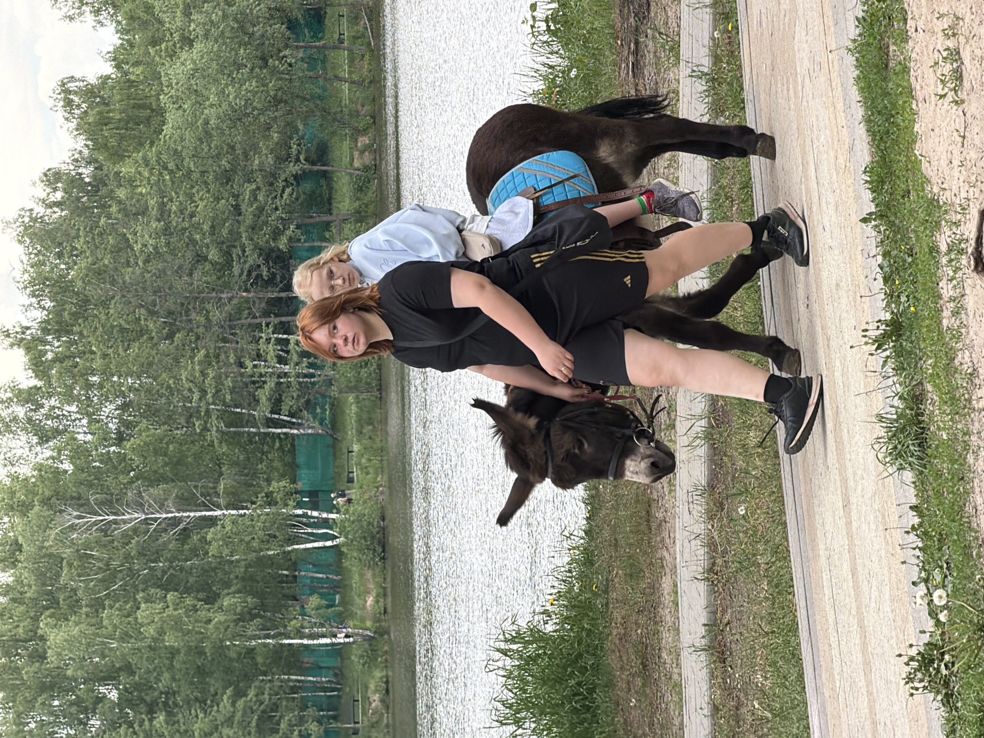 Two young women are riding a donkey on a paved path. The woman in the front is wearing a black shirt and black shorts. The woman in the back is wearing a light blue sweatshirt. The donkey is brown with a light blue saddle. A lake and trees are visible in the background. The location is Malakhovka, Russia.