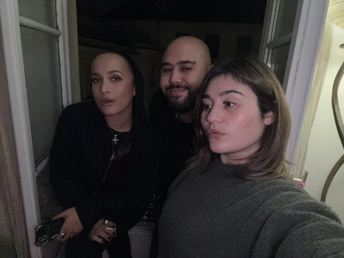 A dimly lit photograph depicts three individuals, two women and one man, positioned within an indoor space featuring an open white casement window, suggesting a nighttime scene in the city of Paris, France. The leftmost individual is a woman with dark hair, wearing a black hooded sweatshirt and a silver cross necklace. She holds a dark-colored smartphone in her left hand, which displays multiple rings on her fingers and dark-painted nails, and gazes directly at the camera. In the center, a bald man with a beard and a dark shirt winks with his right eye while looking towards the camera. The rightmost individual is a woman with medium-length brown hair, clad in a dark green knitted sweater, looking to the right side of the frame with a slightly pouty expression. A portion of an indoor lamp with a patterned shade is visible on the far right of the image.