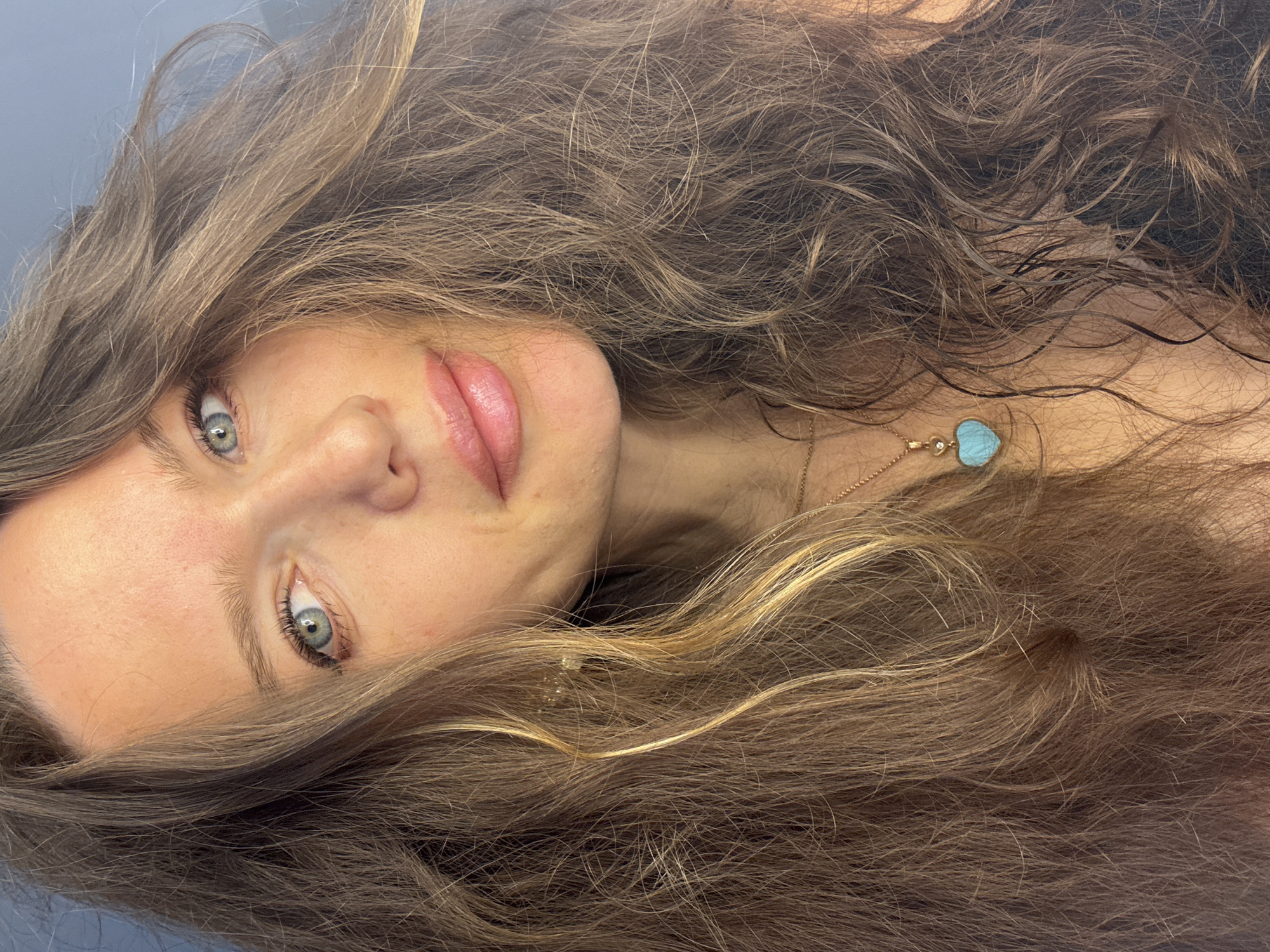 A close-up, slightly tilted portrait of a woman with long, wavy brown hair that is partly obscuring her face. She has light skin, light blue-green eyes, and light pink lips. A gold necklace with a turquoise heart-shaped pendant is visible on her neck. The background is a solid, muted blue-gray color. The location, Khimki, Russia, is not determined from the image.
