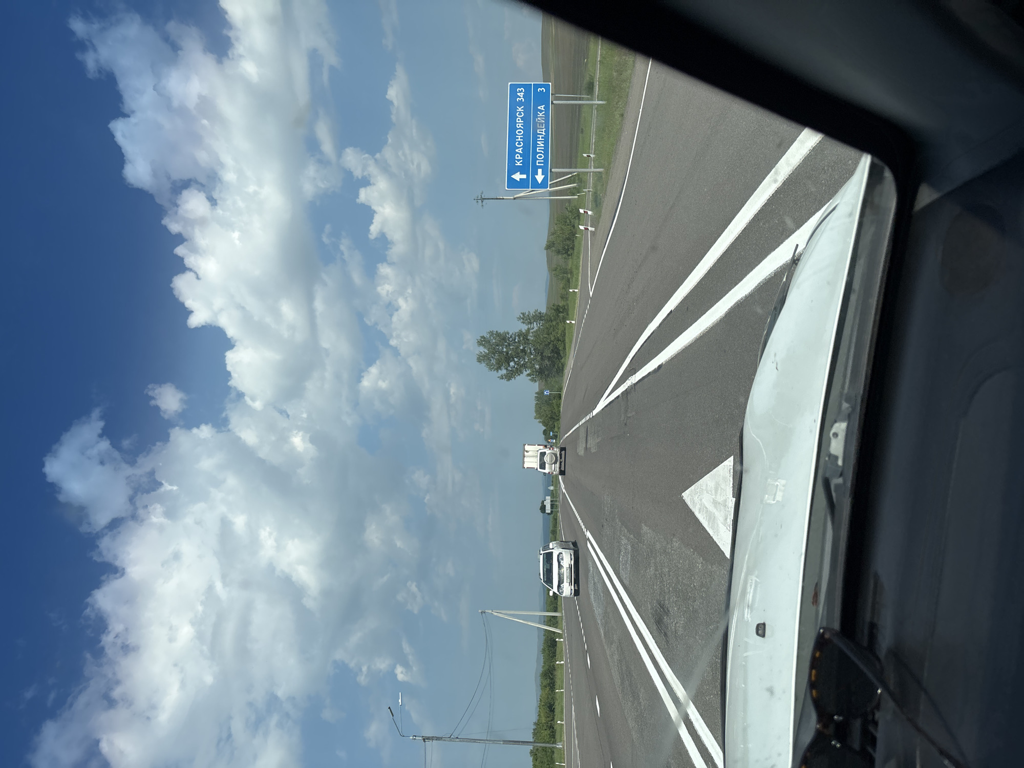The image shows a highway scene from the perspective of a driver. The road is paved with asphalt and has white lane markings. There are several vehicles on the road, including a white car, a truck, and a white SUV. In the background, there are green trees, fields, and a blue sky with white clouds. A blue road sign indicates that Krasnoyarsk is 343 kilometers away and Polindeika is 3 kilometers away to the left. The weather appears to be clear and sunny. The image does not provide specific information about the city of Borodino, Russia.