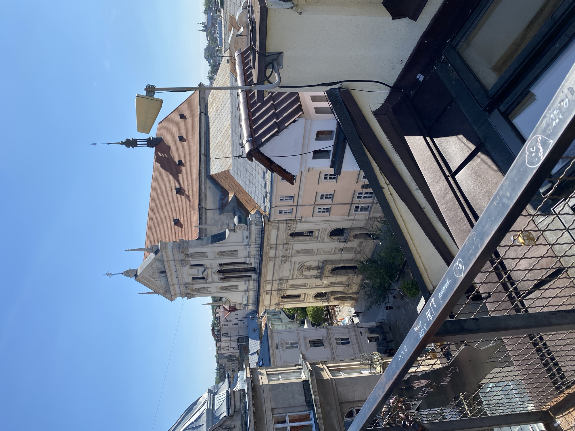 This is an outdoor, high-angle shot of a city street and rooftops in Lviv, Ukraine, on a clear, sunny day. In the background, a large, ornate, light-colored building with a red-tiled roof and a cross on top dominates the view. To its left, a building with a silver dome is partially visible. In the foreground, a metal railing with visible graffiti and padlocks runs across the bottom of the frame. Several other buildings with red and grey tiled roofs are visible, as well as some trees and a street with a few people walking. A light-colored, triangular device is mounted on a pole on one of the rooftops. The overall impression is of an urban landscape with historical architecture.