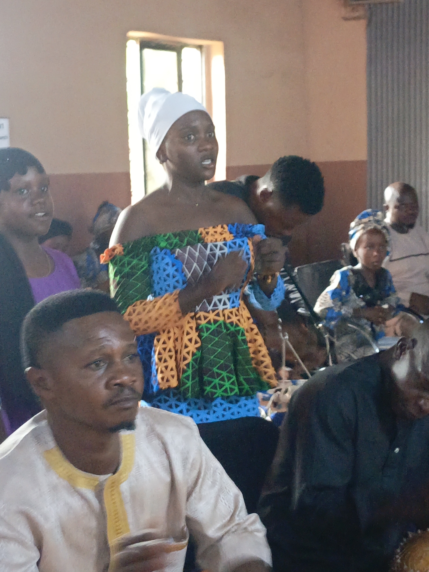 A slightly high-angle, candid shot shows a gathering of people, presumably in Akwanga, Nigeria, based on attire and possible regional context. A woman stands out in the middle ground, wearing an off-the-shoulder dress that features intricate, colorful geometric patterns (blue, green, and orange). Her head is covered with a white headscarf. The woman is speaking or singing, with her mouth open. To her left is another woman in a purple top and a man in a cream-colored traditional shirt with gold trim. In the background, there's a small child wearing a blue patterned headwrap and dress. To the right, another man is visible. The setting seems to be indoors, with a window in the background providing some natural light. There is corrugated metal on the background wall. The other persons wear different clothing and appear to be listening to her. The atmosphere appears to be one of community.