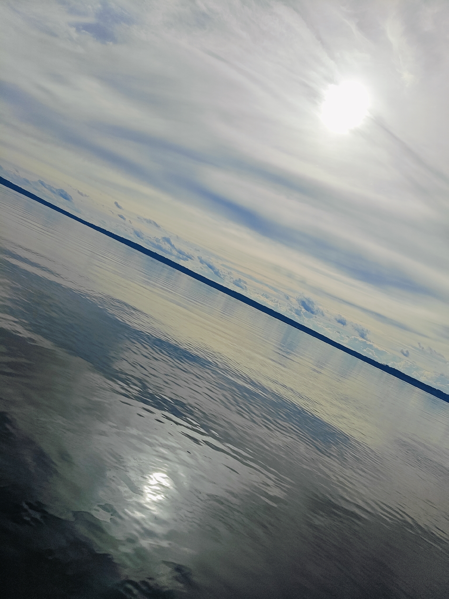 The image shows a diagonal view of a body of water, likely a lake or large river, with a thin dark line representing a distant shoreline or horizon. The sky above is filled with white and blue streaky clouds, with the bright sun visible in the upper right quadrant, casting reflections on the water's surface. The water itself has a rippled texture. The scene is devoid of any people or man-made structures, suggesting a natural, open environment. The location provided, Stepanovo, Russia, indicates a geographical context but is not visually discernible within the image itself.