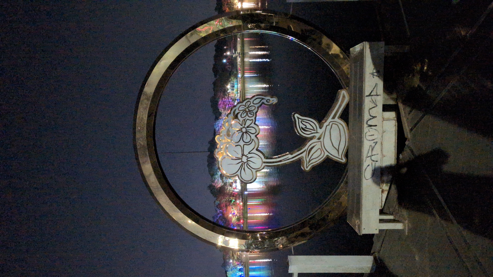 The image depicts a circular metal frame artwork, featuring a stylized floral design, positioned near Xuan Huong Lake in Da Lat, Vietnam. The artwork is likely located on a pedestrian walkway or park area adjacent to the lake. The floral design appears to be cut out of metal with a white or light-colored finish, and the circular frame has a golden or bronze color. The artwork is mounted on a white painted support structure, with some graffiti present on the vertical support section. The background of the frame showcases Xuan Huong Lake, reflecting the city lights, illuminated buildings, and possibly decorative lights along the shoreline. The sky is dark, indicating either night or a very overcast day. The environment suggests an urban park setting, possibly popular with tourists and locals.