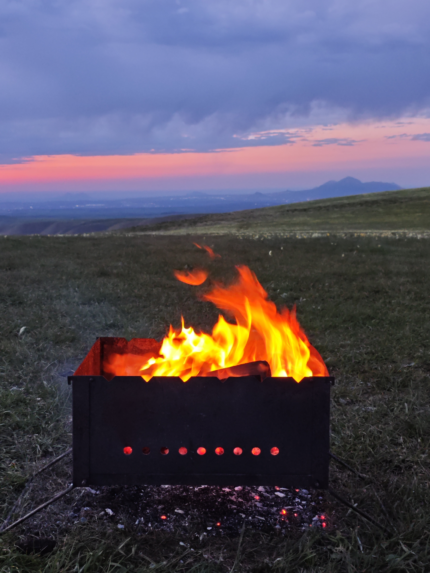 A charcoal grill is burning in a grassy field under a sunset sky. The grill is black and rectangular with a series of holes along the bottom edge. Bright flames and embers are visible inside the grill. The background features a rolling landscape with hints of urban illumination in the distance. The sky is a mix of dark blue and pink hues, indicating a twilight or sunset scene. This photo was taken in the city of Lermontov, Russia.