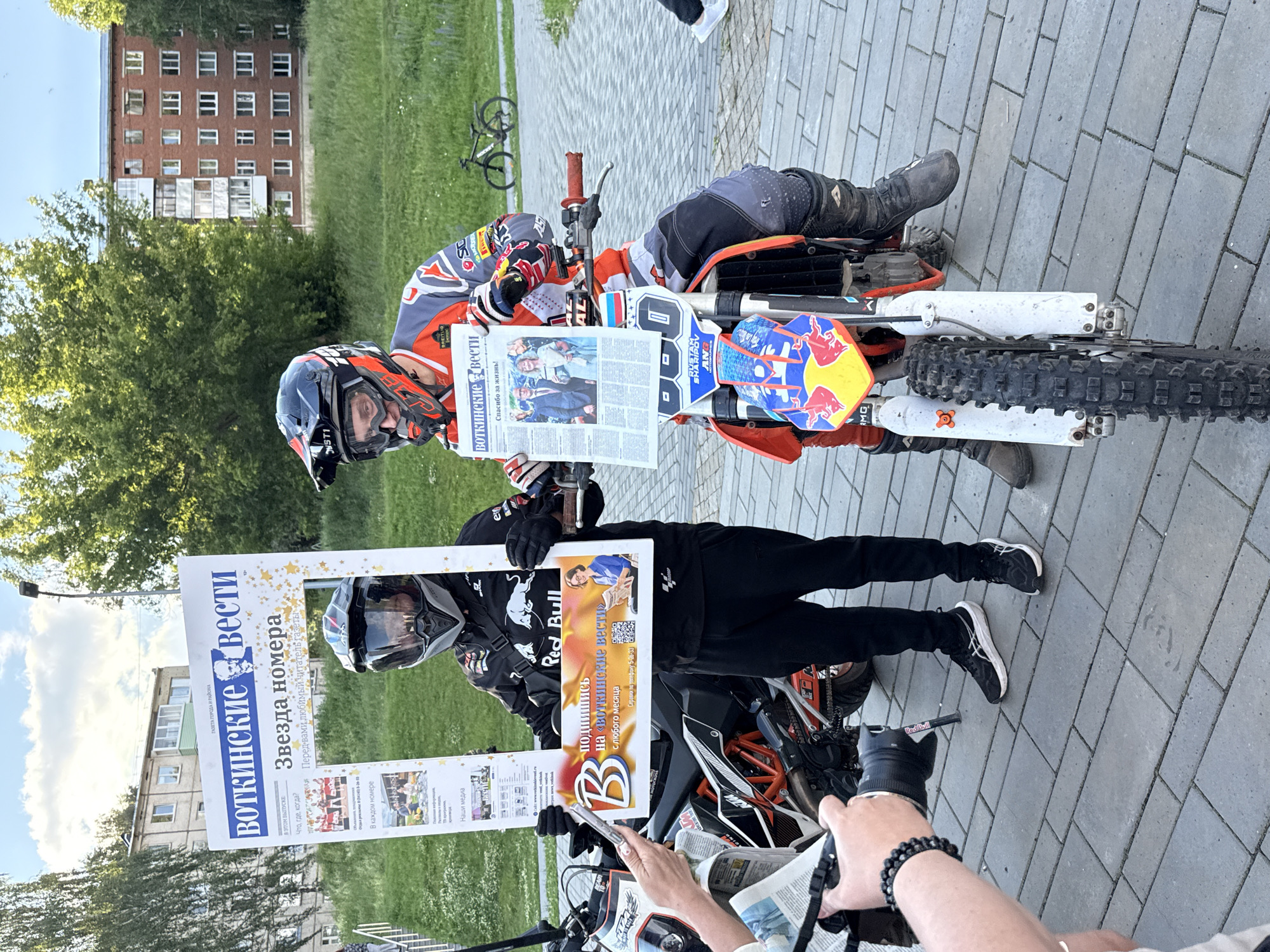 The image captures an outdoor scene in the city of Votkinsk, Russia, likely on a sidewalk area adjacent to a grassy patch and residential buildings. Two individuals, dressed in motorcycle gear, are the central focus. One is standing and holding a large, oversized newspaper prop, while the other is seated on a dirt bike, also holding a newspaper.
The standing individual is wearing a black jacket with "Red Bull" branding and black pants. They are holding a large white frame with text that includes "ВОТКИНСКИЕ ВЕСТИ" (Votkinsk News) and "Звезда номера" (Star of the Issue). Below this, there is a smaller section with text and an image of a woman reading a newspaper.
The individual on the dirt bike is wearing a red, white, and grey racing suit and helmet. The dirt bike itself is predominantly orange and white, with "Red Bull" logos prominently displayed on its front fender and a Russian flag sticker. The rider is holding a standard-sized newspaper, and the front of the bike has a number "0" visible.
In the foreground, a person's arm and hand are visible, holding a camera and seemingly taking a picture of the scene. The ground is paved with grey rectangular bricks. In the background, there are trees, a multi-story brick apartment building, and a lamppost, typical of an urban environment. The lighting suggests it is daytime, with a partly cloudy sky. The overall scene appears to be a promotional event or a photo opportunity involving a local newspaper and motocross enthusiasts.