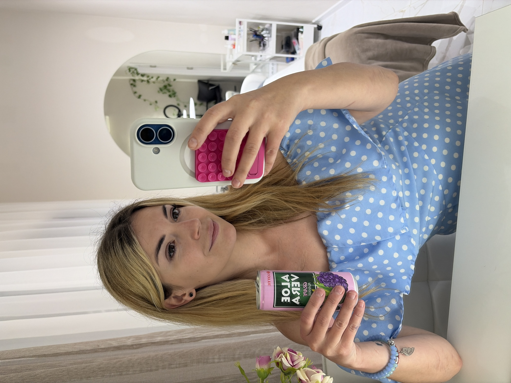 A young woman with blonde hair and dark eyes is captured in a mirror selfie, holding a pink can of "Lotte Aloe Vera Grape" flavored drink. She wears a light blue dress with white polka dots and a light blue beaded bracelet with a silver heart charm on her wrist. Her expression is calm with a slight smile. She holds her phone, which has a white case and a pink pop-socket, in her right hand.
The setting is an indoor room, likely in Budva, Montenegro, characterized by white walls and a large, rounded mirror. The mirror's reflection reveals white shelves stocked with various items and a small green plant. A neutral-toned blanket or throw is also visible in the background. In the foreground, a white surface holds a small bouquet of pink and white roses. The scene is brightly lit, suggesting daytime, with vertical blinds visible through a window, indicating natural light.
