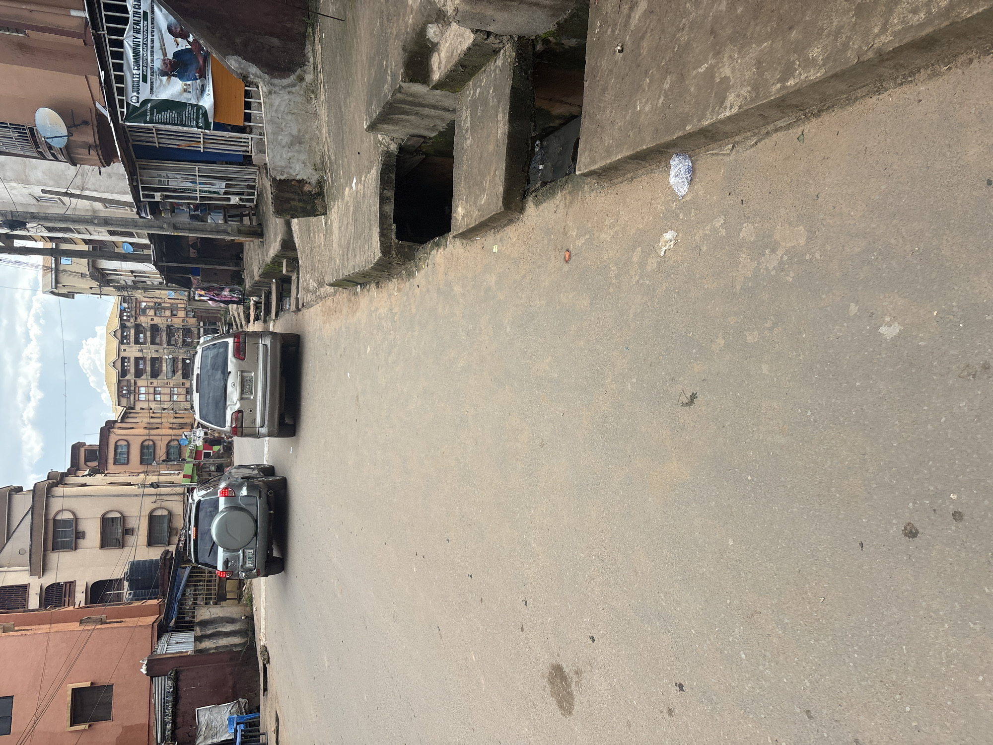 This is a medium-angle, eye-level, outdoor, natural-light photograph of a street in Onitsha, Nigeria. The street appears to be paved with asphalt and shows signs of wear and tear, with some debris and stains present. On the right side of the street, there is an open concrete drainage system with some water and trash visible within it. Several multi-story buildings line the street, some appearing to be residential and others commercial. The architecture is varied, with some buildings having balconies and satellite dishes.
In the foreground, a silver SUV is parked on the left side of the road, facing away from the camera. Further down the street, another silver SUV is also parked, facing the same direction. A banner is hanging on the side of one of the buildings, advertising "Jubilee Community Health Clinic and Dispensary Enterprise." Several people are visible in the background, some walking on the sidewalk and others near the buildings. The sky is overcast with some clouds. The overall impression is of a busy urban environment in Nigeria.