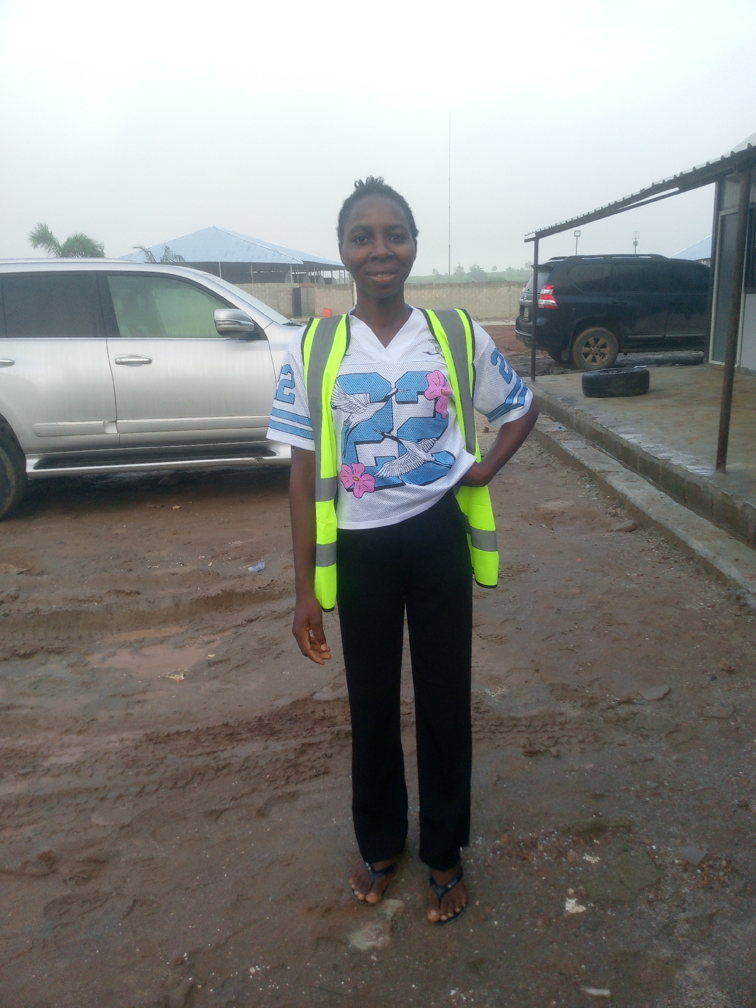 A woman stands in front of a silver SUV and a black SUV in a dirt lot. The woman is wearing black pants, a white t-shirt with a blue and pink graphic of birds and the number 23, and a bright yellow safety vest. She is smiling and has her left hand on her hip. The SUVs are parked on a muddy surface. In the background, there is a building with a metal roof and a concrete wall. The sky is overcast. The location is a street in Indo, Nigeria.