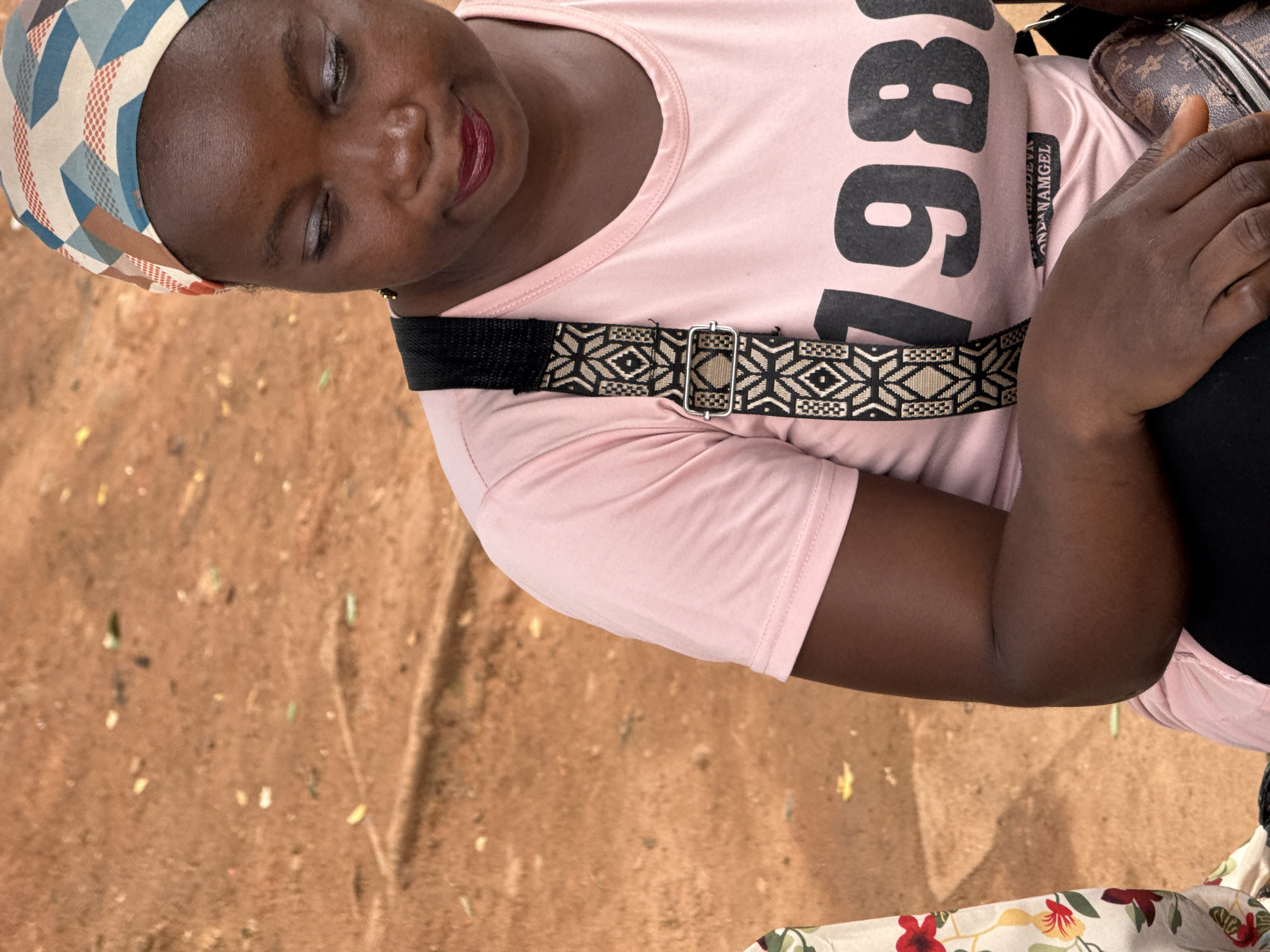 A medium close-up shot shows a Black woman wearing a pink t-shirt with "1981" printed in large black numerals. She has a colorful headwrap and is looking down with her head tilted slightly to the right. Her left arm is visible, resting on her lap, and she is holding a brown patterned bag with her right hand. A strap with a black and white geometric pattern is visible across her chest. The background is a reddish-brown dirt surface with scattered small debris, possibly indicating an outdoor setting in Abuja, Nigeria. The lighting suggests it is daytime.