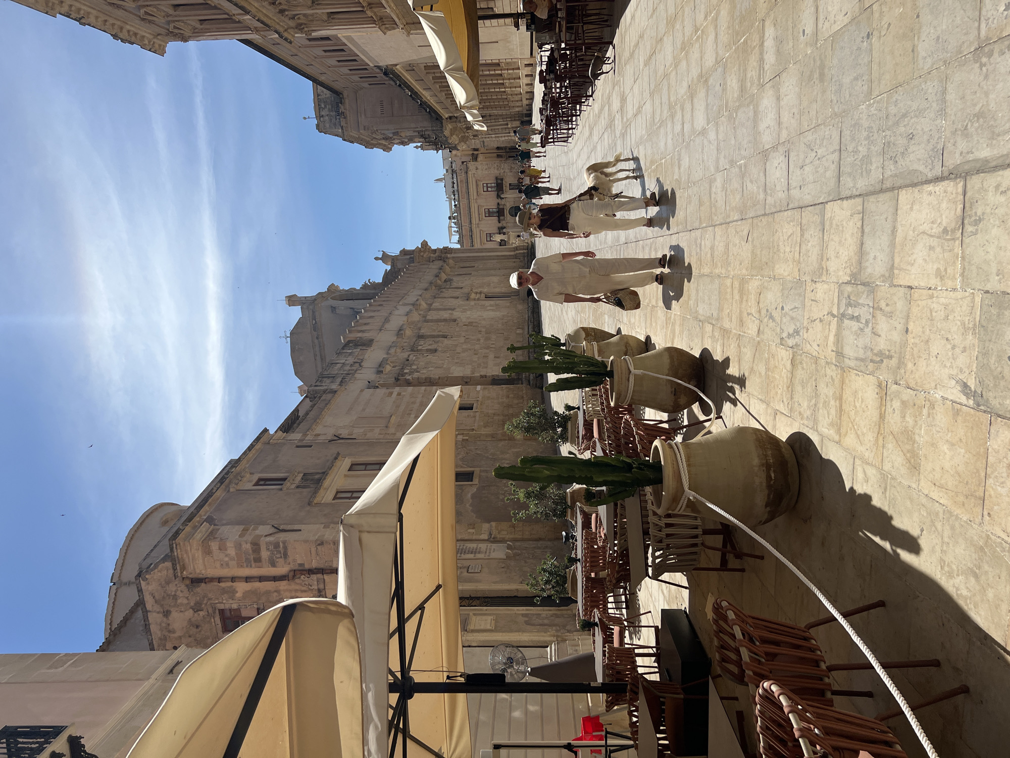 The image depicts a sunlit street scene in Syracuse, Italy. The street is paved with light-colored stone tiles, and flanked by ancient, weathered stone buildings with arched windows and architectural details indicative of Baroque style. On the left, an outdoor cafe is set up with several tables and chairs covered by large, cream-colored umbrellas. Large terracotta pots with tall cacti are strategically placed along the edge of the cafe area.
In the center of the street, a couple is walking a dog. The man, wearing a light-colored cap and a matching linen outfit, carries a straw bag. The woman beside him wears a wide-brimmed hat and a brown top with white pants. They are walking towards the viewer, with the dog, a light-colored sighthound breed, walking on a leash beside them. Further down the street, other people are visible walking, and more outdoor seating for a cafe is arranged. The sky is a clear blue with a few wispy clouds, and the bright sun casts distinct shadows across the pavement. The overall atmosphere suggests a warm, pleasant day in a historic European city.