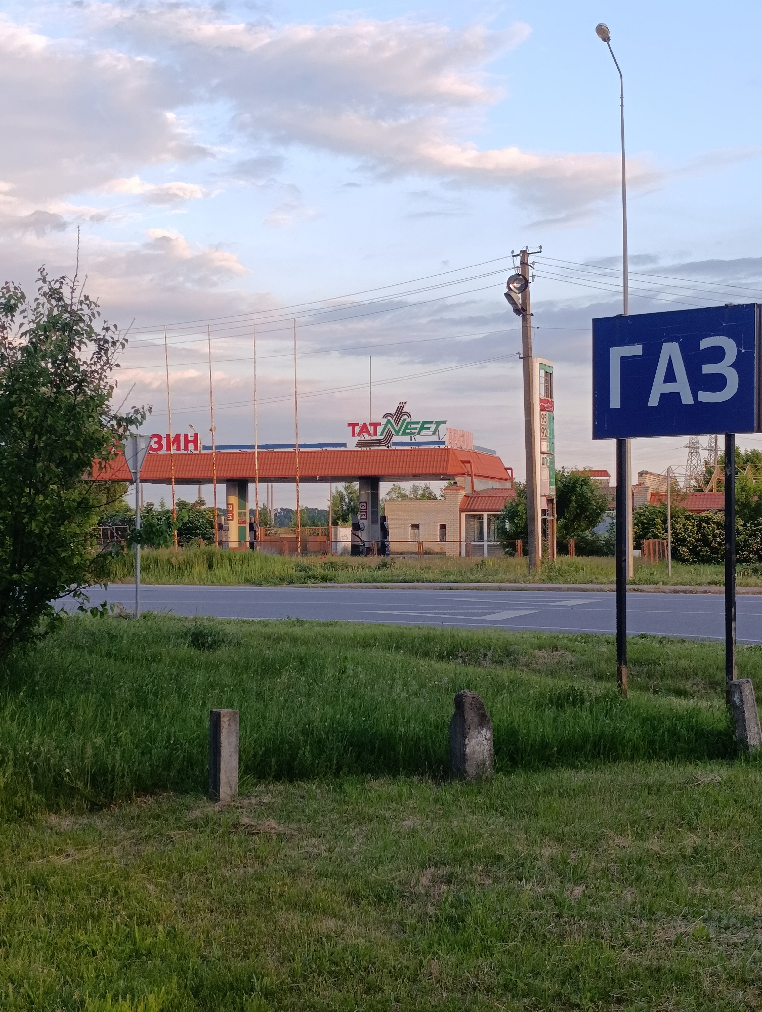 A wide shot depicts a gas station and a road under a partly cloudy sky, likely during late afternoon or early evening. In the foreground, a vibrant green grassy area stretches across the bottom of the image, featuring several small, weathered concrete posts. A paved asphalt road with white markings runs horizontally across the midground.
Beyond the road, the gas station comes into view. It features a building with a distinctive red-tiled canopy roof. On the canopy, parts of Cyrillic text are visible: "ТАТНЕФТЬ" (TATNEFT), indicating the brand, and "ЗИН", likely part of "БЕНЗИН" (BENZIN), meaning gasoline. In front of the canopy, several tall, rusty metal poles or supports stand. To the right of the main building, a tall gas price sign displays "95", "92", and "АТГ" (likely referring to Autogas/LPG).
To the far right, a large blue road sign with prominent white Cyrillic letters reads "ГАЗ" (GAZ), typically indicating a natural gas or LPG filling point. A tall street light pole stands behind this sign, and a utility pole with power lines is visible further left. On the left side of the frame, a leafy green tree partially obstructs the view of the gas station. The background consists of more trees and some indistinct buildings. There are no visible people or vehicles, suggesting a quiet moment at the station in Krasnohorivka, Ukraine.