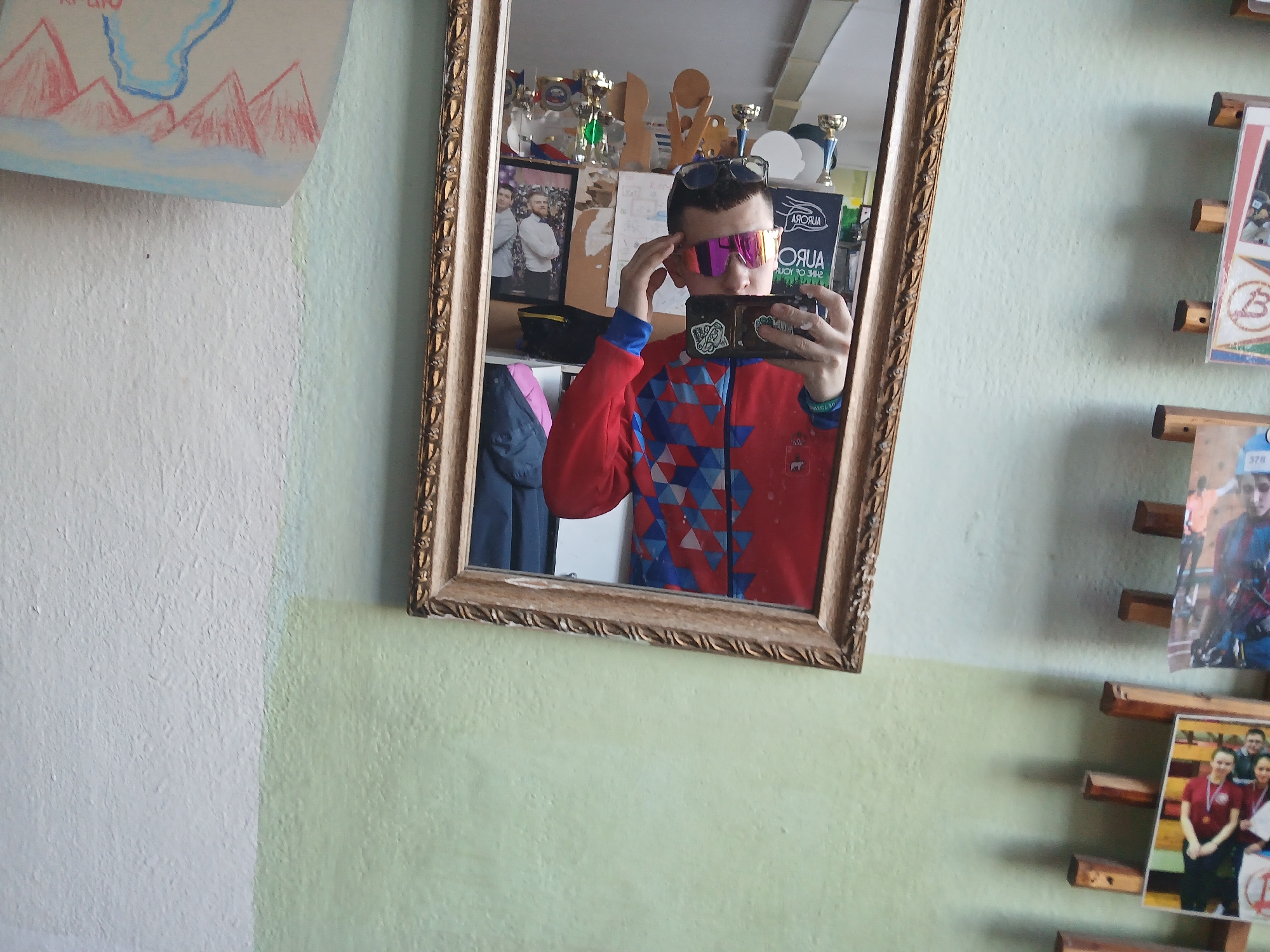 The image shows a selfie of a person in a mirror. The person is wearing a red jacket with blue triangle patterns and sunglasses with pink lenses. They are holding a phone with stickers on it, taking a picture in the mirror.
Behind the person in the mirror, there are several trophies, a framed picture of two men, and a book. To the left of the mirror, there is a white and green wall with a drawing of mountains hanging. To the right of the mirror, there is a wall with pictures attached to a wooden slat system.
Given the presence of items associated with the Russian sports team "Aurora" and the trophies, the location may be related to sports activities. Based on previous questions and knowledge, Berezniki, Russia, is relevant.