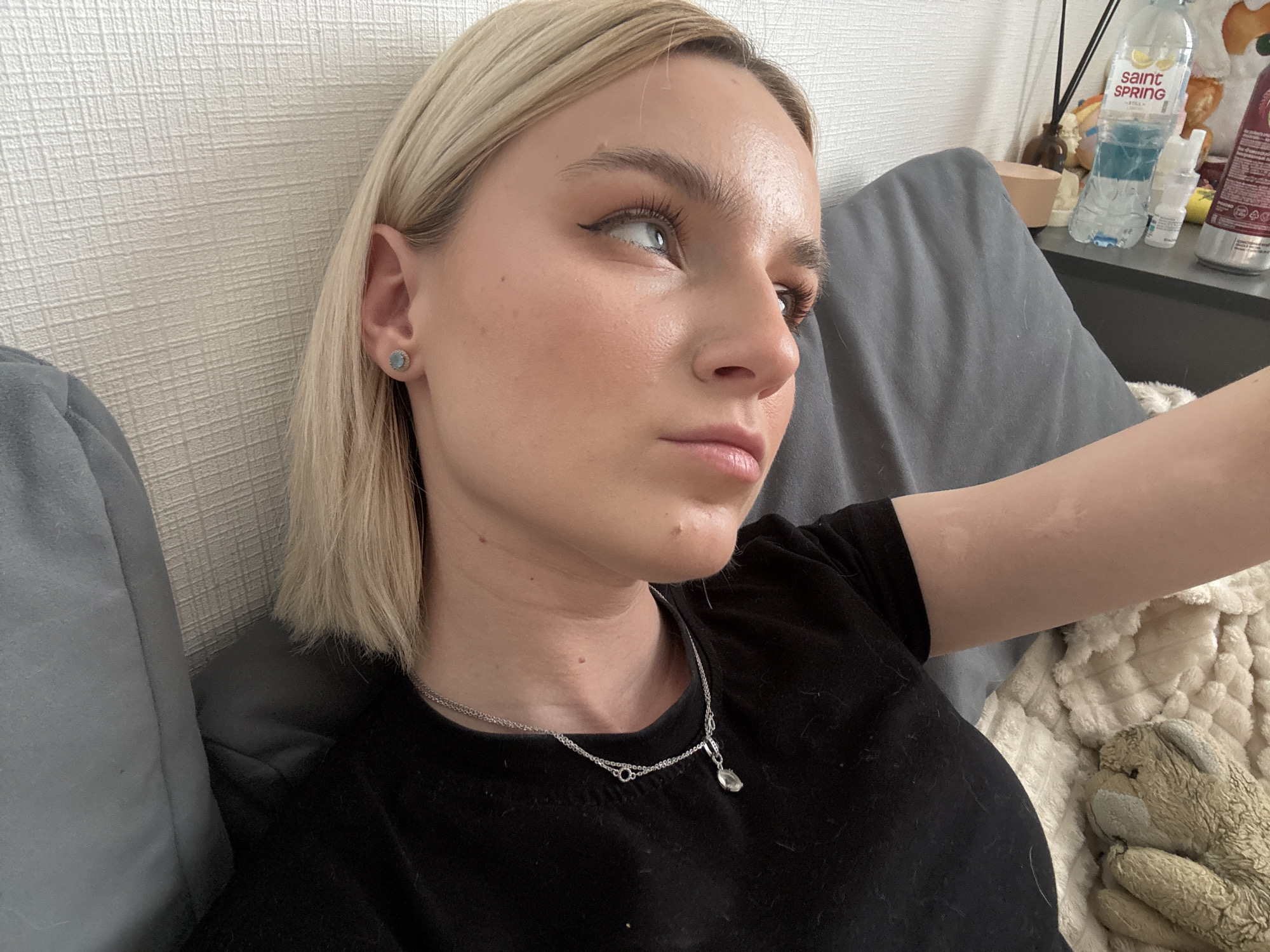 A young woman with blonde, shoulder-length hair is lying on a bed or couch. She is wearing a black t-shirt and a silver necklace with a pendant. She has subtle makeup on, including winged eyeliner. She is looking off to the right side of the frame with a pensive expression. A gray pillow is behind her head. To the right of her arm is a fluffy blanket and a brown teddy bear. In the background, there's a table or shelf with a bottle of "Saint Spring" water, some small bottles, and decorative items. The location is not identifiable as Vsevolozhsk, Russia from the visual information provided.