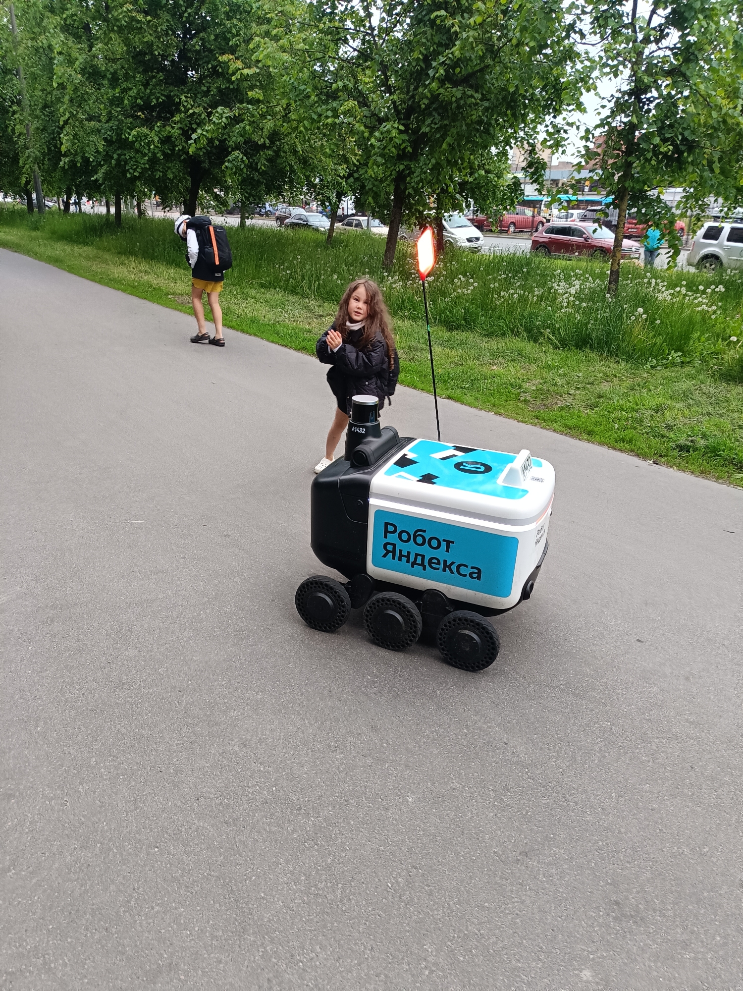 The image captures a scene in a city, featuring a young girl interacting with a Yandex delivery robot on a paved path alongside a grassy area with trees. The robot is white and black, with the words "Робот Яндекса" (Robot Yandex) in Cyrillic script on its side, and is equipped with six wheels and a vertical red light. The girl is dark-haired and wears a black jacket, white socks, and white shoes. She stands close to the robot with her hands clasped in front of her. In the background, a person with a backpack walks away from the camera on the same path, while cars and other urban elements are visible in the distance. The environment consists of trees, grass, and a street with cars, suggesting an urban or suburban setting.