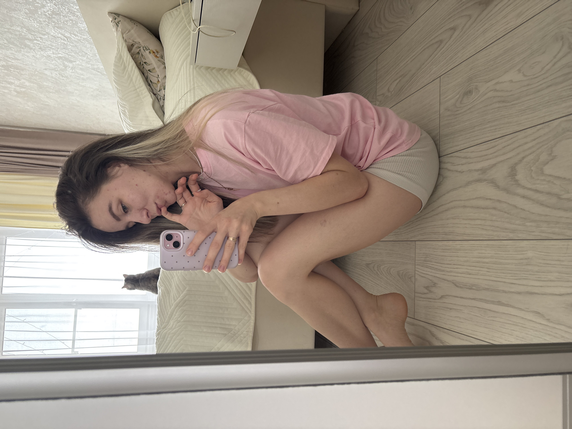 A young woman with dark hair and blonde highlights is seated on a light-colored wooden floor. She is wearing a pink t-shirt and light-colored shorts. She is holding a lavender-colored phone and appears to be taking a selfie in a mirror. Behind her, a cat is visible on a white bed near a window. A beige nightstand is also visible. The reflection suggests an indoor setting. The city of Noviy, Russia, cannot be independently verified based on the image.