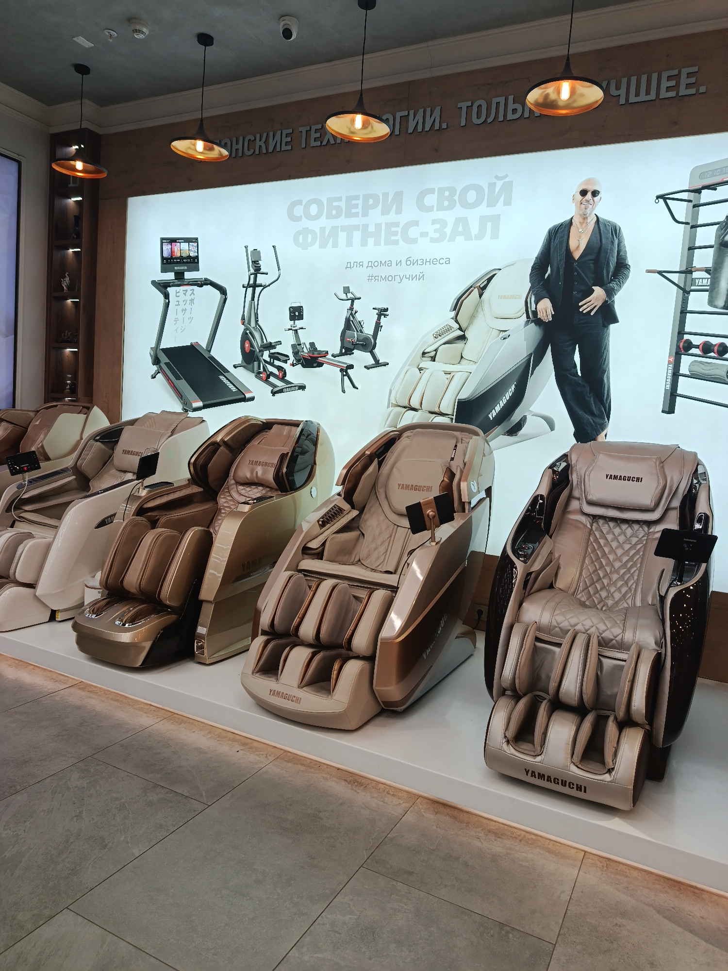 The image shows a showroom, likely located in Moscow, Russia, featuring a display of massage chairs. Five chairs of different models and colors, all bearing the "Yamaguchi" brand name, are arranged in a row on a white platform. Behind the chairs, a large promotional poster showcases a man in a suit and sunglasses leaning on one of the massage chairs. The poster also displays fitness equipment such as a treadmill, an elliptical machine, a rowing machine, and a weight rack, along with the slogan "СОБЕРИ СВОЙ ФИТНЕС-ЗАЛ" (Assemble your gym) and the hashtag "#ямогучий" (#IamPowerful). The upper part of the poster is partially obscured by a wooden frame and three hanging pendant lights. The floor is covered with gray rectangular tiles. A display shelf with decorative objects is visible on the left side of the image. A security camera is installed on the ceiling.