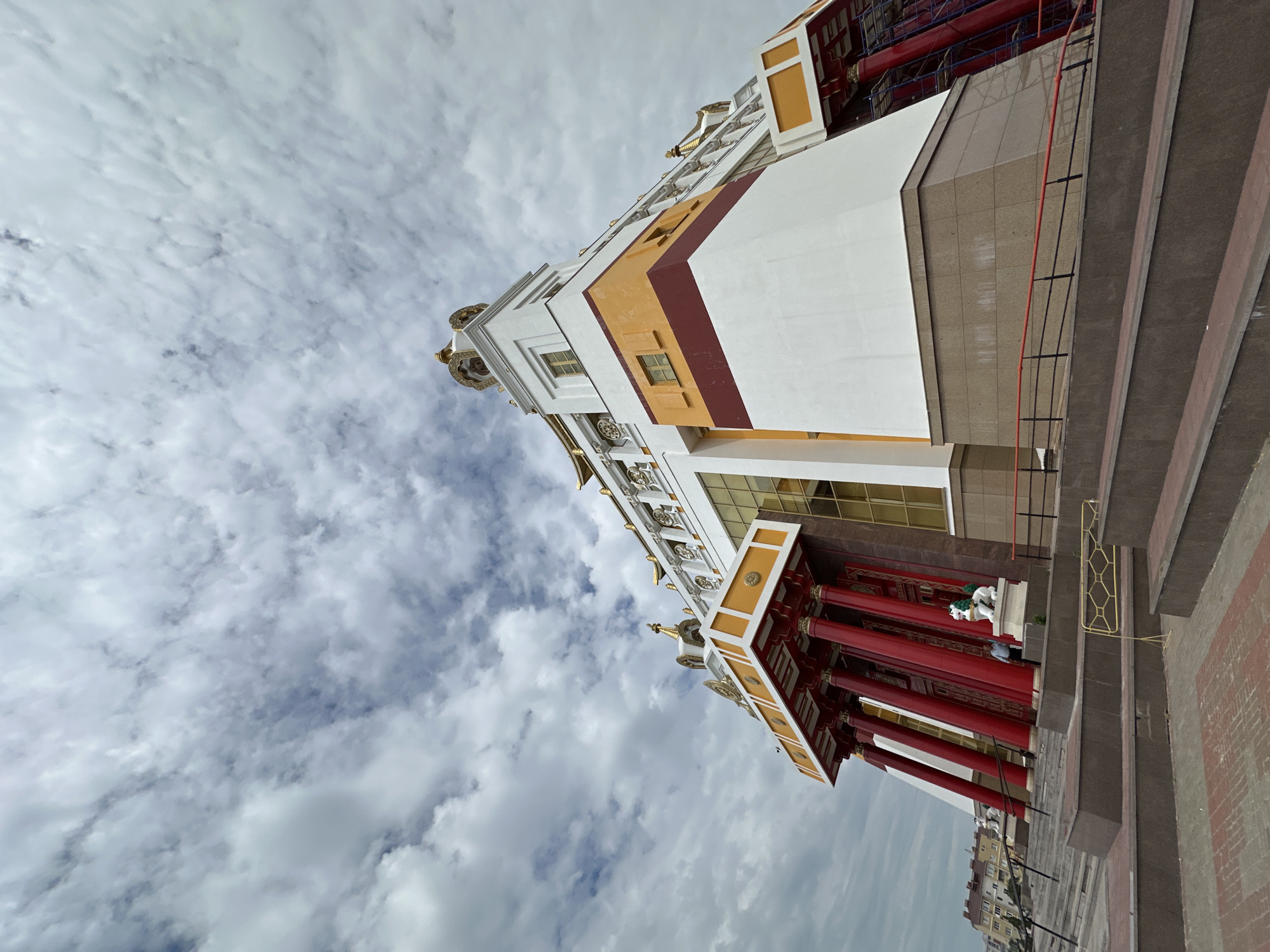 A low-angle, wide shot captures the exterior of the Golden Abode of Buddha Shakyamuni, a Buddhist temple in Elista, Russia. The building, characterized by its white, orange, and maroon facade, features ornate golden accents on its roof and a grand entrance with tall, red pillars. The temple is situated against a backdrop of a cloudy sky, with a few residential buildings visible in the distance. In front of the temple, a man stands near a statue of a lion. The foreground is dominated by a series of stone steps leading up to the entrance.