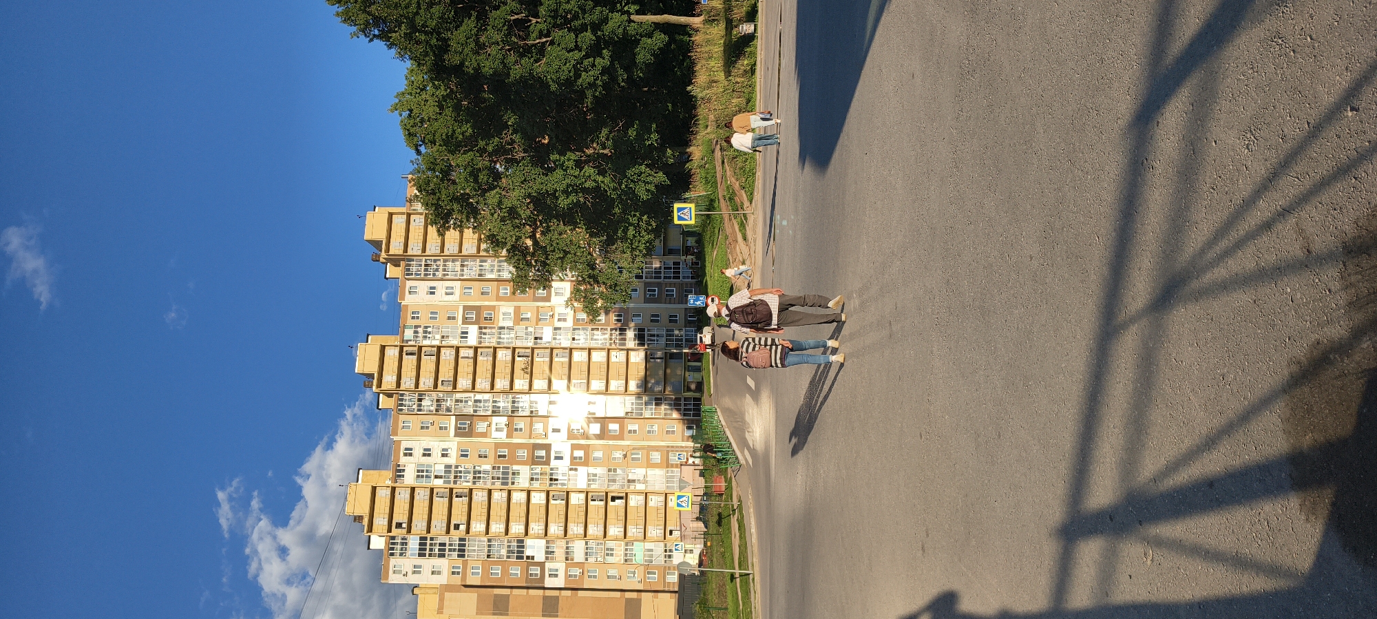 In an outdoor urban setting under a clear blue sky with scattered clouds, a large, multi-story yellow apartment building dominates the background. The building has many windows and balconies. To the right of the building, a large, mature tree with dense green foliage is visible. In the foreground, a paved road or intersection is present, with pedestrian crossing markings. Two people, a woman and a man, are walking away from the camera across the road. The woman is wearing a striped shirt, blue jeans, and a backpack. The man is wearing a plaid shirt, dark pants, and a backpack. Several other people are visible in the distance, walking or standing. Yellow pedestrian crossing signs are positioned along the roadside. The scene is illuminated by sunlight, casting long shadows on the road.