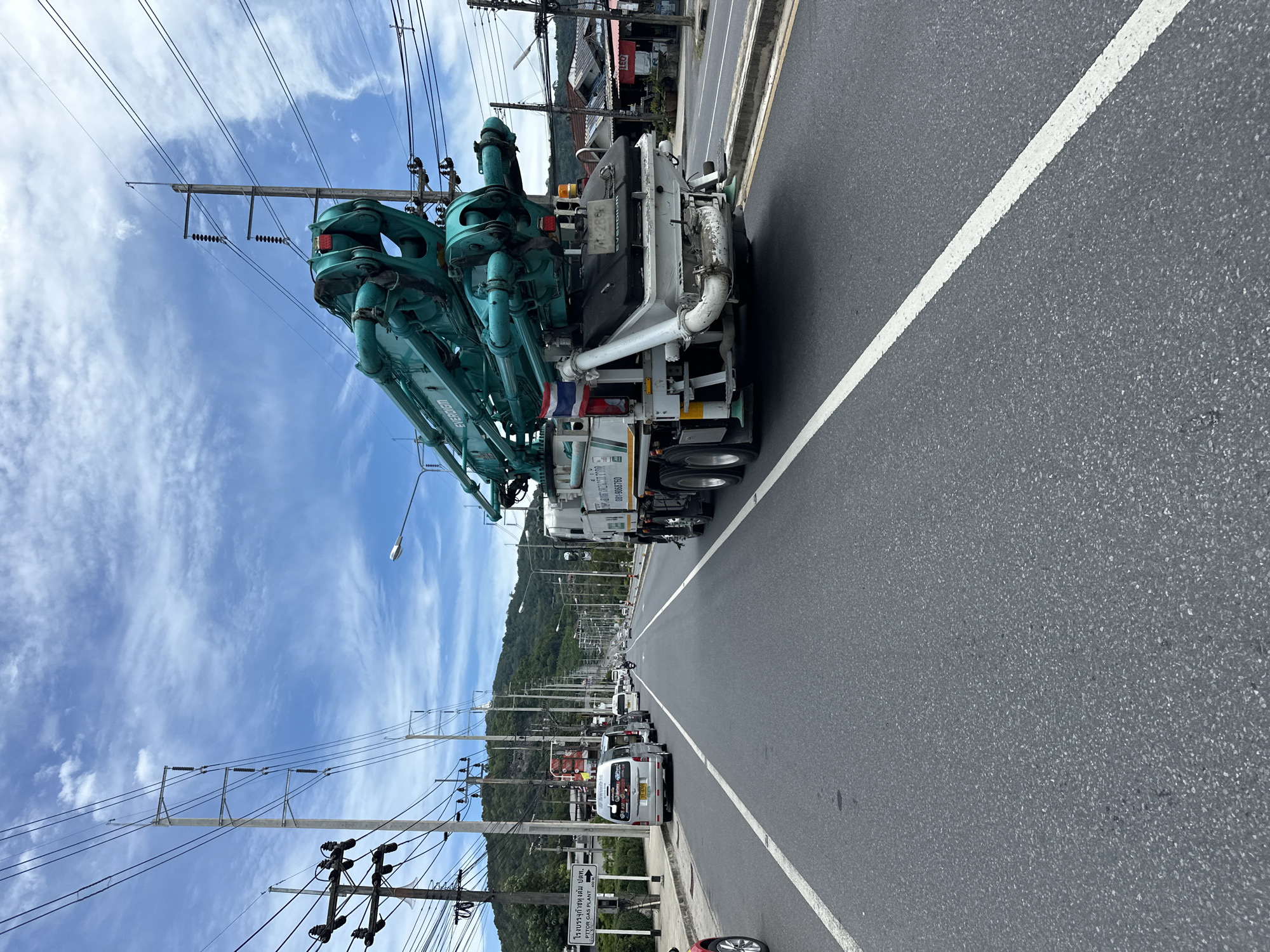 The image captures a view from the perspective of a vehicle traveling on a paved road. A large, teal-colored concrete pump truck dominates the right side of the frame. The truck has multiple articulated boom sections and a white chassis. A Thai flag is visible on the side of the truck. In the distance, the road stretches towards a mountainous landscape under a cloudy blue sky. Numerous utility poles with power lines line the sides of the road. Several other vehicles, including vans and motorcycles, are also present on the road. The scene appears to be in an urban or semi-urban area with buildings visible on the right side of the road. A sign with Thai text is visible on the left side of the road. The road has white lane markings.