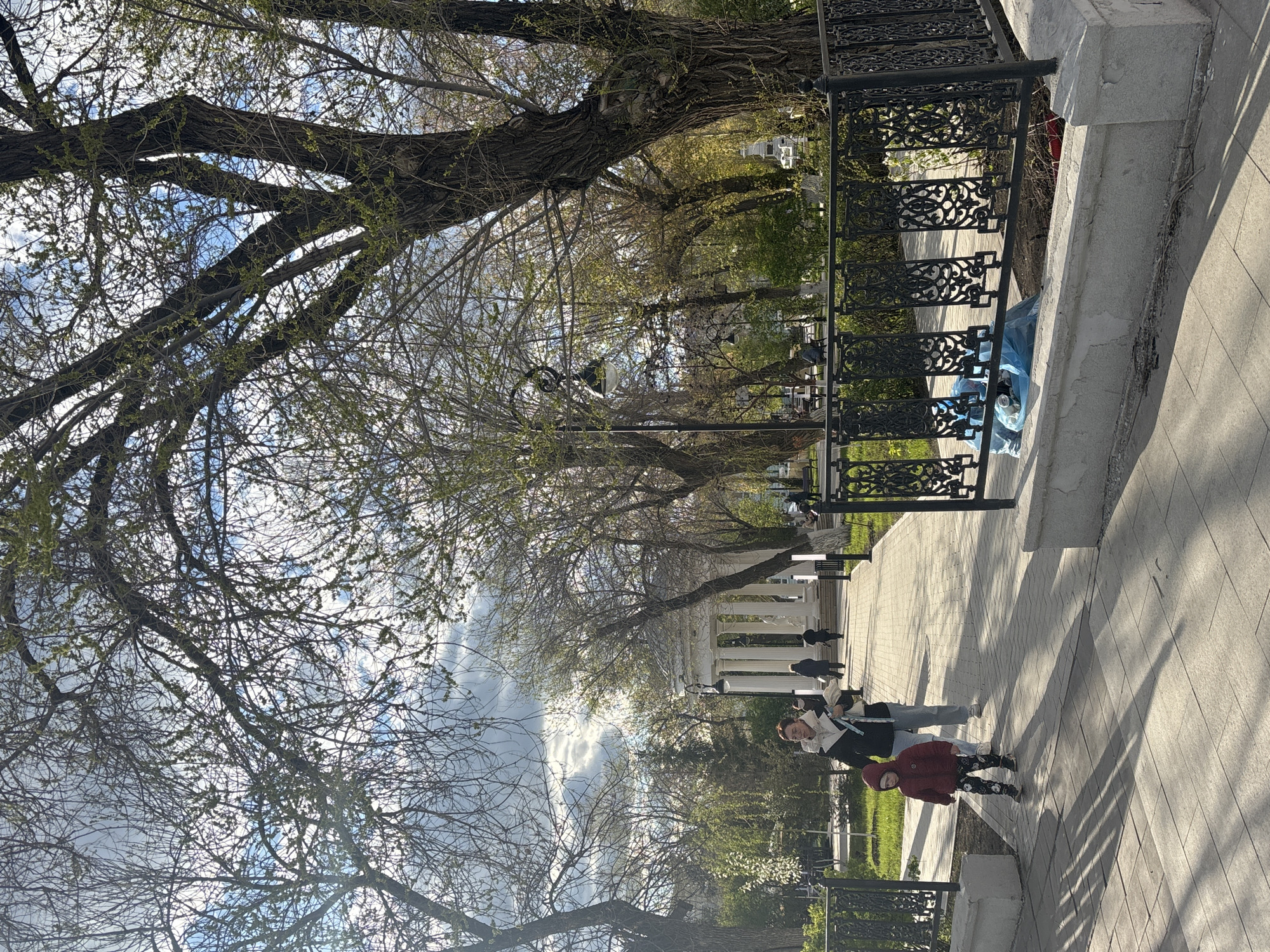 The image depicts a park in Orenburg, Russia. A paved walkway lined with wrought iron fencing and mature trees is visible. The trees have bare branches and emerging leaves. A white colonnade structure is in the background. Three people are walking on the pathway: an adult with a light jacket and a black bag, an adult with a dark jacket, and a child with a red jacket and patterned leggings. Litter in a blue bag is behind the fence.