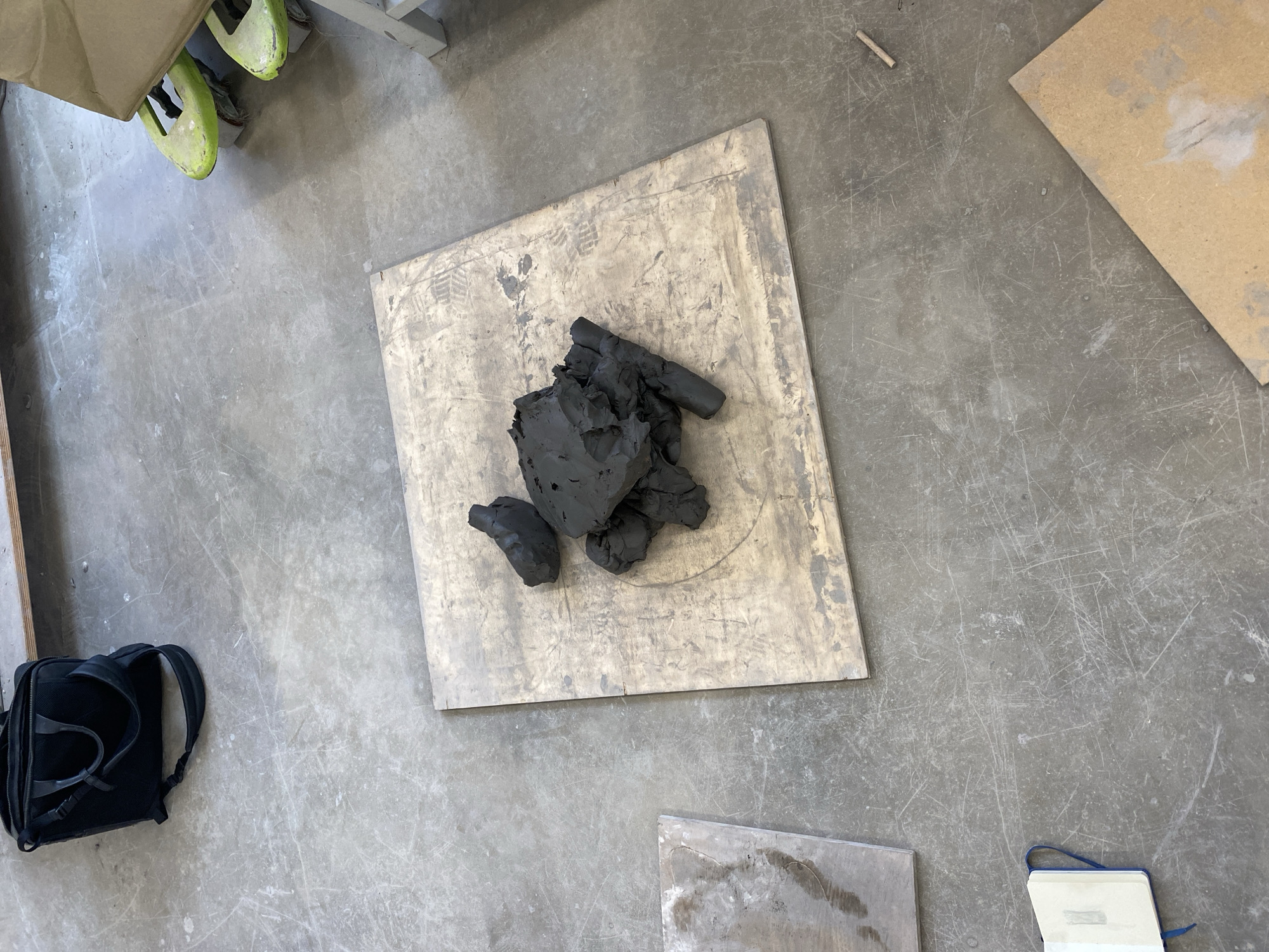 An overhead shot captures a section of a concrete workshop or studio floor, likely in Saint-Ouen-sur-Seine, France. The central focus is a mound of dark, possibly black, modeling clay piled on a rectangular light-colored wooden board. The board itself is worn, featuring circular indentations, smudges, and visible footprints, suggesting it is a well-used surface for creative work.
The concrete floor displays numerous scratches, scuffs, and dust, indicative of a functional and active workspace. In the upper left quadrant, the bright lime-green soles or parts of two shoes or sandals are visible, partially obscured, hinting at someone standing nearby. A black backpack with its straps visible rests on the floor in the lower left corner.
Other items include a smaller, lighter-colored wooden board in the upper right portion of the frame, and a slender, short wooden stick situated nearby. In the lower right corner, a small, open notebook with a blue binding is present. The scene depicts a momentary pause in artistic or crafting activity, with raw materials and personal items laid out in a practical, unadorned setting. No discernible text is visible on any objects within the frame.