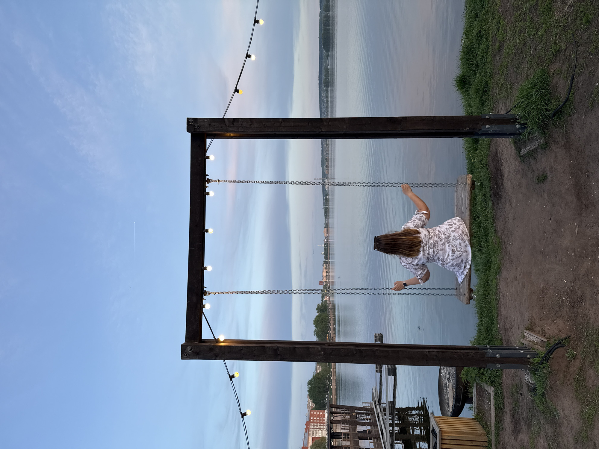 A woman in a patterned dress sits on a swing facing a body of water. The swing is suspended from a dark wood frame adorned with string lights. Beyond the water, the cityscape of Votkinsk, Russia is visible, including buildings and greenery. The foreground also includes grass and dirt. The sky transitions from blue to lighter hues, suggesting either sunrise or sunset.