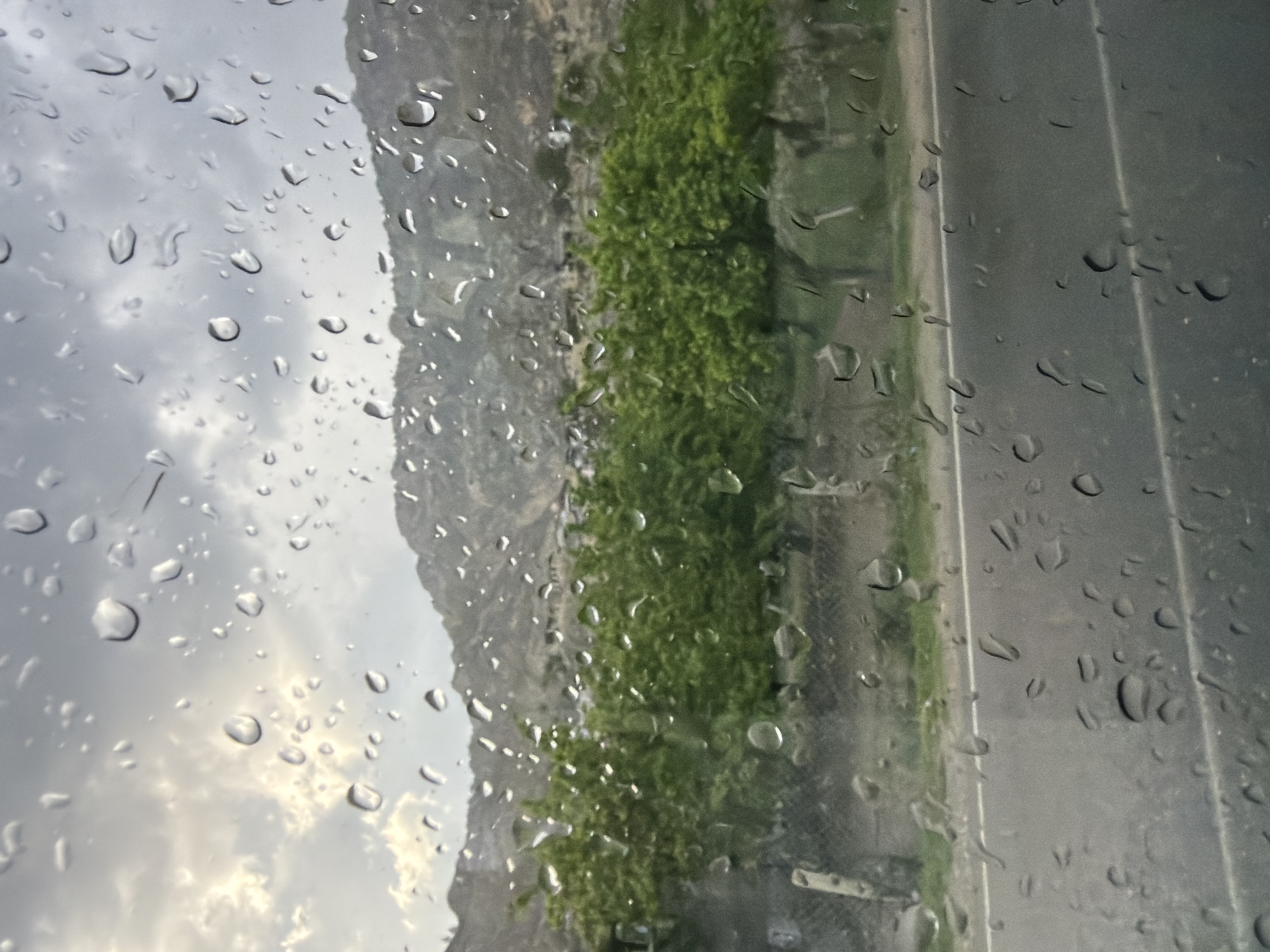 The image shows a view through a rain-streaked window. Outside, there is a roadside with a strip of green vegetation separating it from a rocky hillside. The sky is overcast.