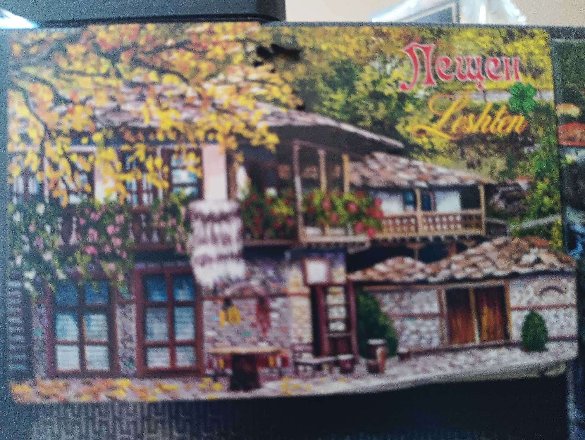 This is a postcard featuring a picturesque scene of a traditional Bulgarian house in the village of Leshten. The house has a distinctive architectural style, with stone walls and a tiled roof. It is adorned with colorful flowers, and the surrounding trees are in full autumn foliage, with leaves in shades of yellow and red. Text on the postcard reads "Лещен Leshten" in red and yellow font. The image captures a serene and rustic atmosphere, typical of Bulgarian rural landscapes. The village of Leshten is located in the Garmen Municipality in Blagoevgrad Province, southwestern Bulgaria, and is known for its preserved Revival-era architecture. It is not located in the city of Karnobat, Bulgaria.