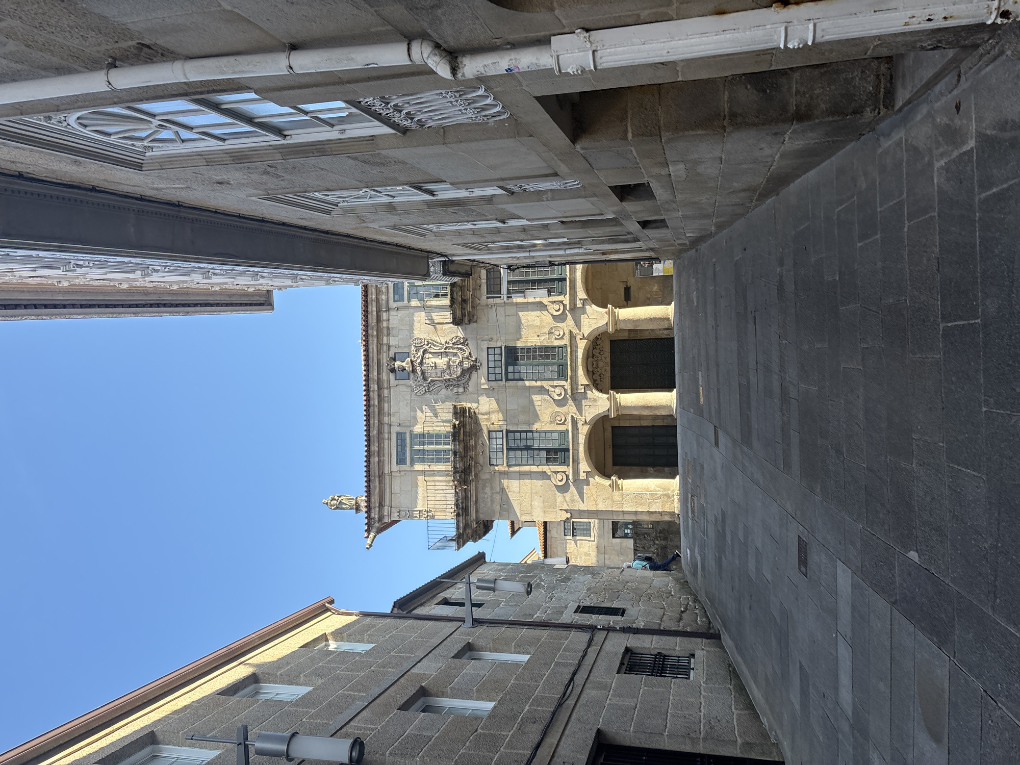 The image depicts a narrow, sloped cobblestone street in the historic center of Pontevedra, Spain. The street is flanked by stone buildings on both sides. On the left, a building with a light-colored stone facade and several windows is visible. On the right, a more ornate stone building with arched doorways and a balcony adorned with a coat of arms and a statue on the roof is prominent. A person, wearing a blue shirt and dark pants, is walking up the street away from the camera. The sky is clear and blue. The architecture suggests a European, likely Spanish, urban setting, consistent with Pontevedra's old town.