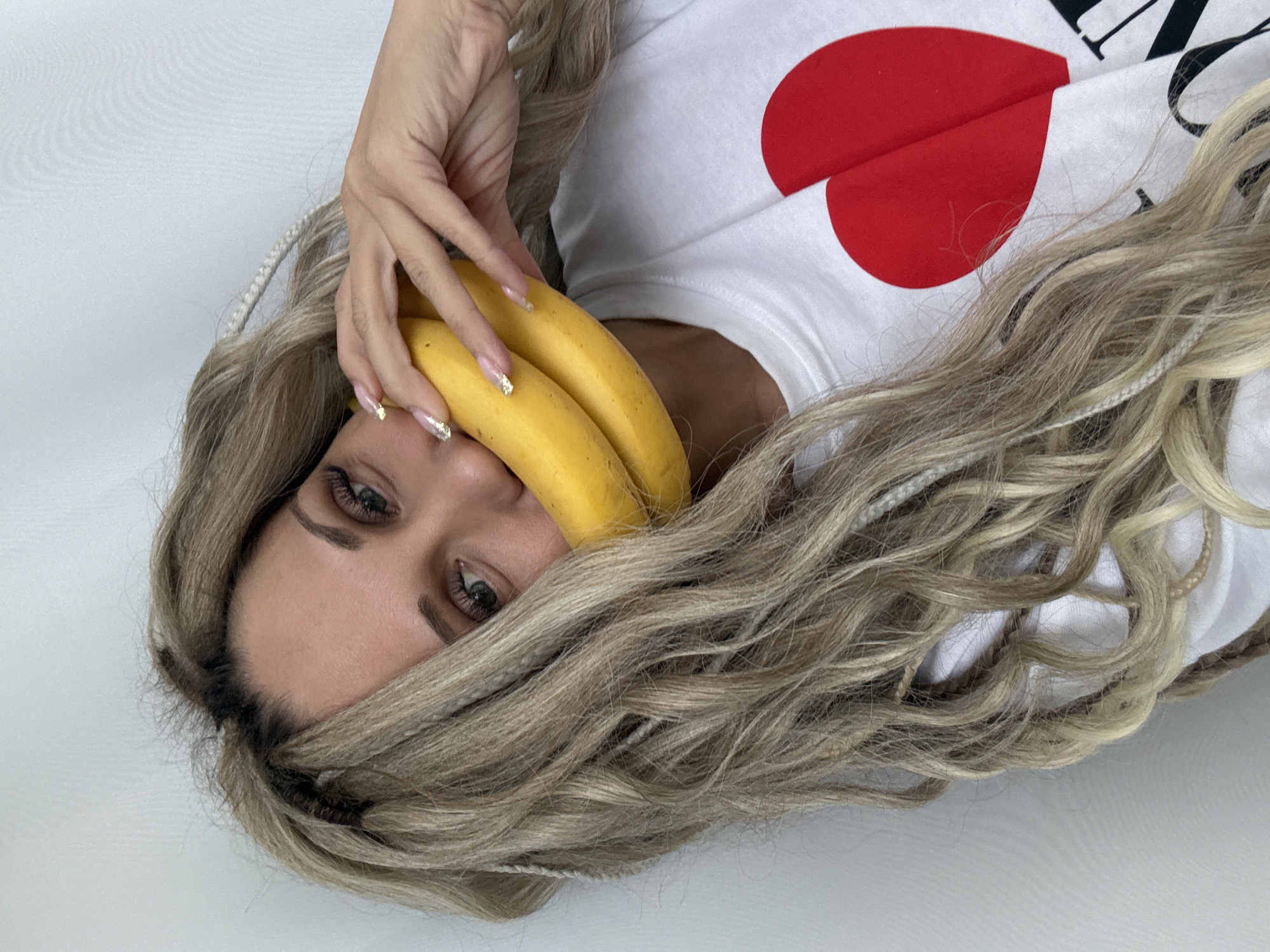 A woman is lying on a white surface. Her head is tilted towards the upper left corner of the frame. She has long, wavy blonde hair with braids that frames her face. She is holding two yellow bananas near her mouth with her left hand. The woman's eyes are visible, and she has light skin and brown eyebrows. She is wearing a white t-shirt with a red heart graphic on the chest. The location is Rusanovo, Russia.