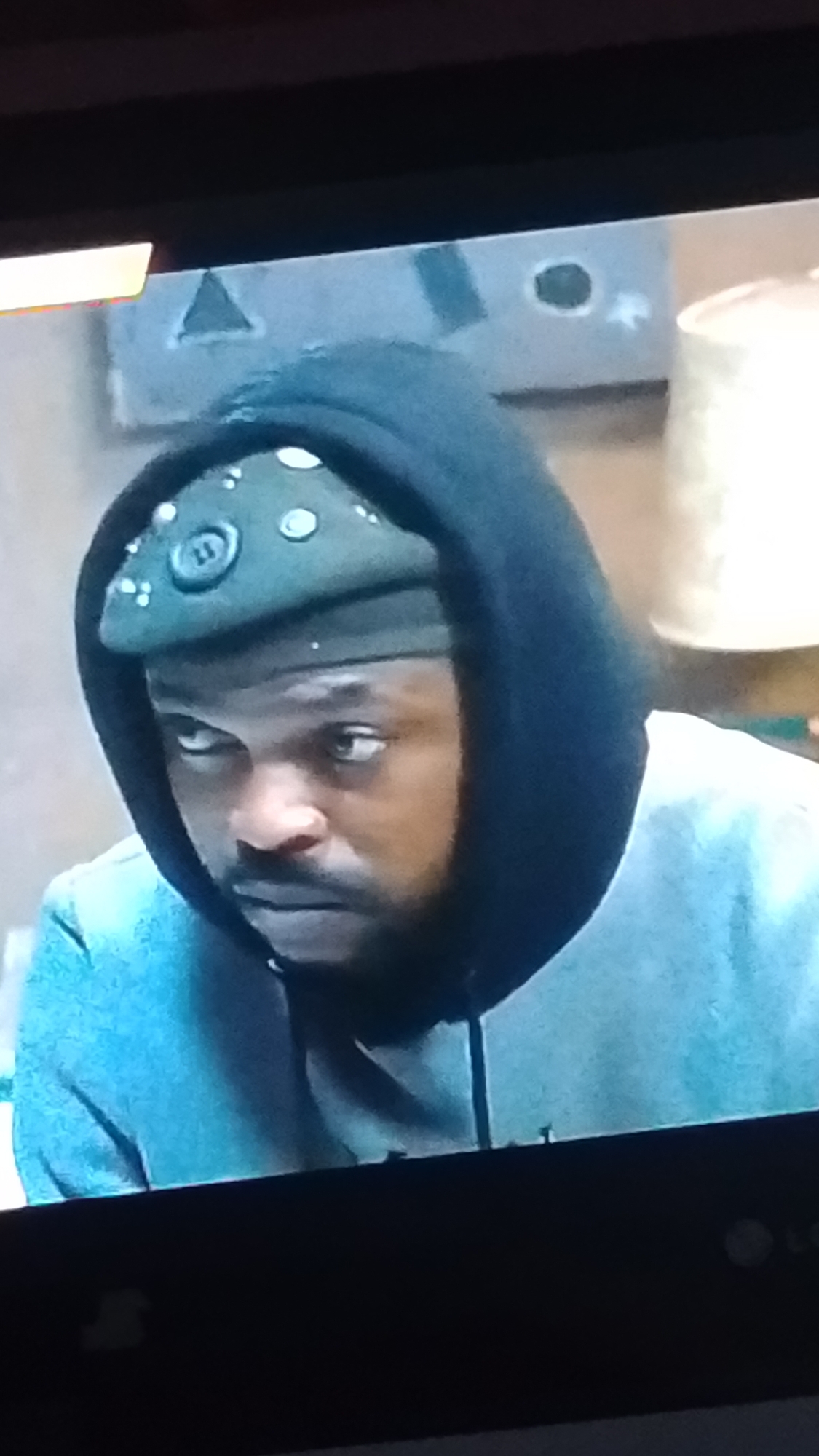 In a setting within Lokoja, Nigeria, a close-up image captures a Black male, appearing to be in his 20s or 30s, with a dark beard and mustache. He is depicted wearing a dark blue or black hooded sweatshirt with the hood pulled up, partially obscuring his ears and neck. Beneath the hood, a dark green or olive-colored cap or bandana is visible, adorned with multiple light-colored, possibly metallic, circular studs or embellishments across its surface. A prominent circular button or badge with the numeral "11" clearly marked is affixed to the front center of this headwear. His gaze is directed intensely towards his left (viewer's right), conveying a serious expression. The man wears a lighter blue or teal garment beneath his hooded sweatshirt, visible around his chest and arms. The background suggests an indoor environment, with a light-colored wall featuring abstract shapes, including a triangular motif on the upper left. A section of a cylindrical, light-colored object, possibly a lamp or lampshade, is visible on the upper right. The entire scene is presented as a photograph taken of a digital screen, indicated by the visible dark bezel and part of an "LG" logo at the bottom right.