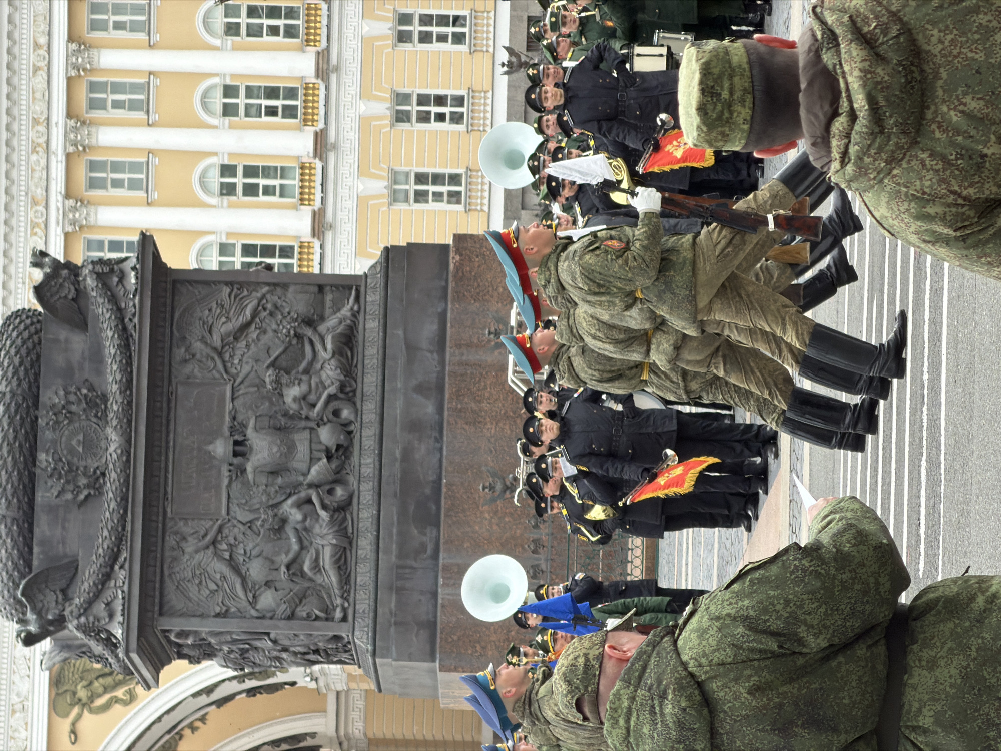 This image depicts a formal military parade or ceremony taking place in a grand urban setting. The perspective is from behind and slightly above a few soldiers in camouflage uniforms, looking towards a marching formation.
In the foreground, on the left, stands the base of a large, dark grey monument or column, adorned with intricate sculptural reliefs depicting figures, eagles, and symbolic elements. The details on this monument are rich and elaborate.
To the right and further back, numerous military personnel are arranged in formations. Soldiers in the immediate foreground wear camouflage uniforms with green and brown patterns, some sporting blue-brimmed hats. They are equipped with rifles featuring wooden stocks and wear tall, polished black boots. Beyond them, a military band is visible, with members in dark blue or black uniforms and hats. They carry various brass instruments, including a prominent white-belled sousaphone, and drums. One soldier holds a red banner decorated with yellow symbols.
In the background, on the far right, a magnificent yellow building with numerous windows, both arched and rectangular, and classical columns, lines the square. This architectural style, combined with the presence of the ornate monument, strongly indicates the location is Palace Square in Saint Petersburg, Russia, with the building likely being the General Staff Building and the monument being the Alexander Column.
The ground is paved, showing clear white lines used for parade formations. The event appears to be occurring on a clear, daylight day. The overall mood conveyed is one of discipline, solemnity, and formality inherent in a military parade. No discernible text is visible on uniforms or banners.