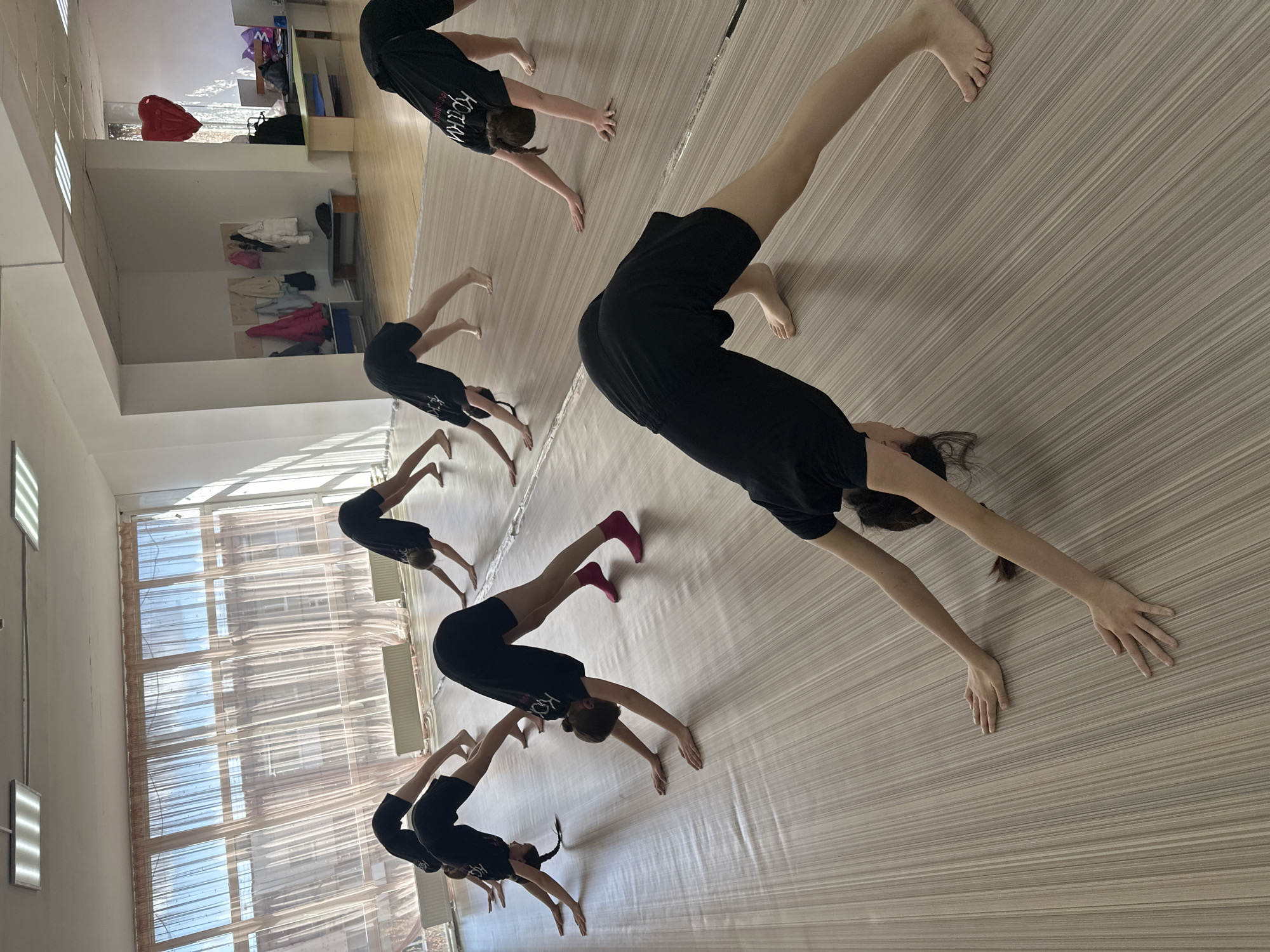 The image depicts an indoor training session in what appears to be a studio or practice space, located in Votkinsk, Russia. Several young individuals, primarily girls, are engaged in a synchronized stretching or conditioning exercise. They are all dressed in matching black t-shirts and shorts, with one participant notably wearing bright pink socks. Their posture involves bending forward with their hands on the floor, backs arched, and legs extended, resembling a downward-facing dog or a preparatory stretch.
The room is brightly lit, with natural light pouring in through large windows on the right side of the frame, which are partially obscured by sheer, light-colored curtains. The clear sky visible through the windows indicates it is daytime. Additional illumination comes from rectangular fluorescent light fixtures on the ceiling.
In the background on the left, shelving units or cubbies hold various items, including folded clothing like jackets and shirts. A distinctive red heart-shaped object, possibly a balloon or decoration, hangs near one of these shelves. A light-colored bench or low table is also visible. The floor is light-toned with a linear pattern, suitable for physical activities.
Visible text on the back of the black t-shirts is in white Cyrillic letters, clearly showing "КОТЧАН", which likely represents the name of the studio or a team. The individuals appear focused and concentrated on their exercise routine.