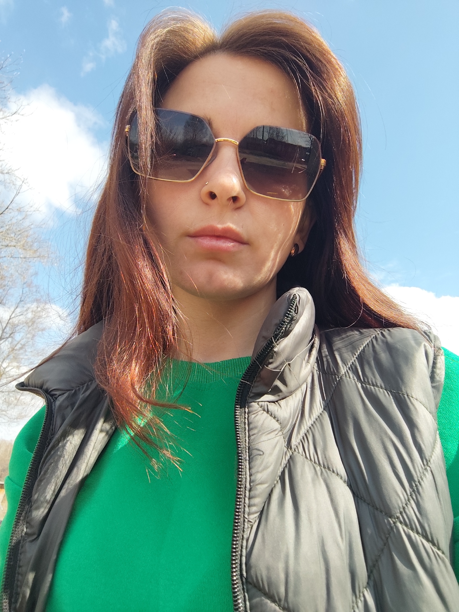 A close-up, upward-facing portrait captures a woman outdoors in Фоки, Russia, likely during a sunny day. She is centered in the frame, wearing dark, oversized square-shaped sunglasses with gold-toned frames. A small metallic stud is visible as a nose piercing on her left nostril. Her long, reddish-brown hair, some strands catching the sunlight, frames her face and falls over her shoulders. She wears a bright green long-sleeved top, partially covered by a dark gray or black quilted puffer vest, which is zipped up.
The woman's expression is neutral, with her lips slightly parted. The bright blue sky, speckled with white clouds, dominates the upper background, indicating clear weather. Bare tree branches are subtly visible in the distant background on the left, suggesting a season like late winter or early spring. Strong, direct sunlight illuminates the scene, casting shadows on the woman's face from the sunglasses and highlighting her hair and the textures of her clothing, particularly the quilted pattern of her vest. No visible text is present in the image.