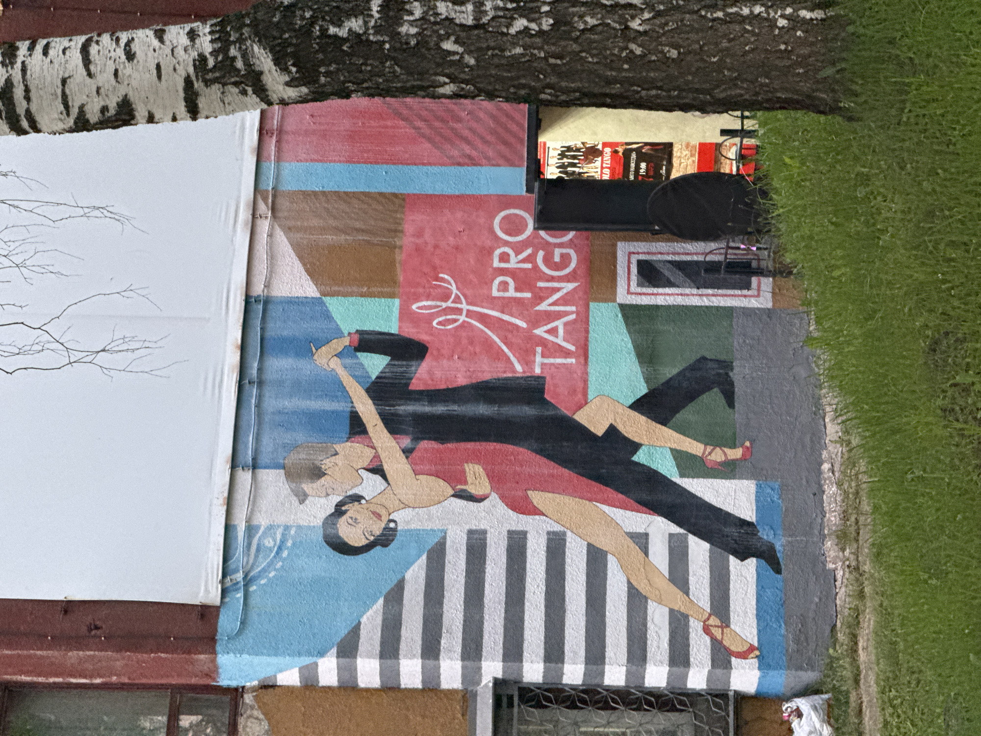 The image shows a mural painted on the exterior wall of a building, depicting a couple dancing the tango. The mural is vibrant and colorful, with the dancers in dynamic poses. The woman is wearing a red dress with a high slit, and the man is in a black suit. The background of the mural features abstract geometric patterns and stripes. To the right of the mural, there is a black speaker and a poster advertising a tango event. A portion of a birch tree trunk is visible in the foreground, along with green grass. The building appears to be weathered, with some signs of wear and tear. A white plastic bag is visible on the ground near the base of the wall. The overall setting suggests an outdoor urban environment.
This mural is located on the building at the intersection of Komsomolsky Prospekt and Pushkinskaya Street in Izhevsk, Russia. It is a well-known landmark in the city, often referred to as the "Tango Mural." The building itself is a multi-story structure with windows and architectural details consistent with Soviet-era architecture. The mural is a popular spot for photos and a symbol of the city's cultural life. The poster on display indicates a tango event, reinforcing the theme of the mural. The presence of the speaker suggests that this is an active location, possibly a venue for performances or events.