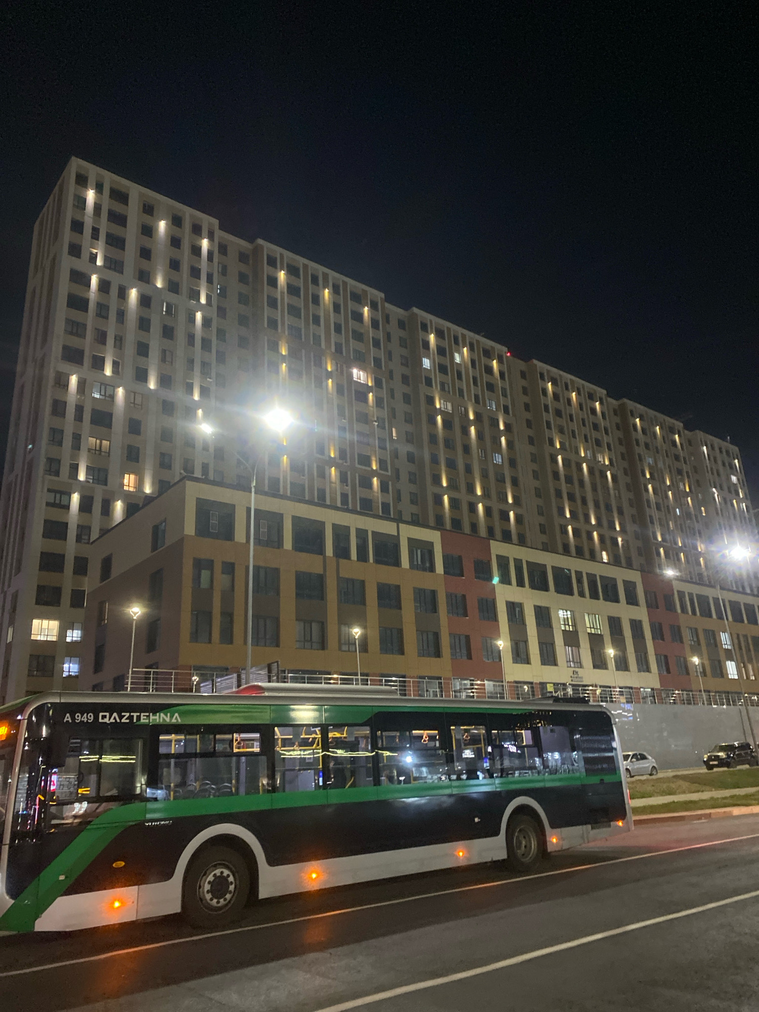 This image depicts a nighttime view of a modern, multi-story apartment building in Astana, Kazakhstan. The building features a light-colored facade with numerous windows, many of which are illuminated, suggesting occupancy. The lower levels of the building are accented with sections of colored panels in shades of brown, orange, and red. In the foreground, a green and black public bus is driving on a road. The bus has text on its side that reads "A 949 QAZTEHNA." Streetlights are visible in front of the building, and a few cars are parked or driving in the background. The sky is dark, indicating it is nighttime.