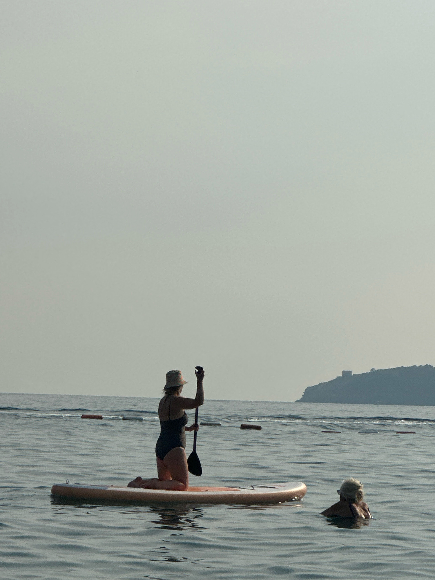 In the city of Boreti, Montenegro, two individuals are visible on the water. In the foreground, a person is kneeling on a paddleboard, holding a paddle. They are wearing a swimsuit and a hat. To the right, another person is partially submerged in the water, with their head and shoulders visible. The paddleboard is positioned horizontally in the frame. In the background, a hazy coastline with a distant tower can be observed, with buoys marking a boundary in the water. The sky is overcast and the water appears calm.