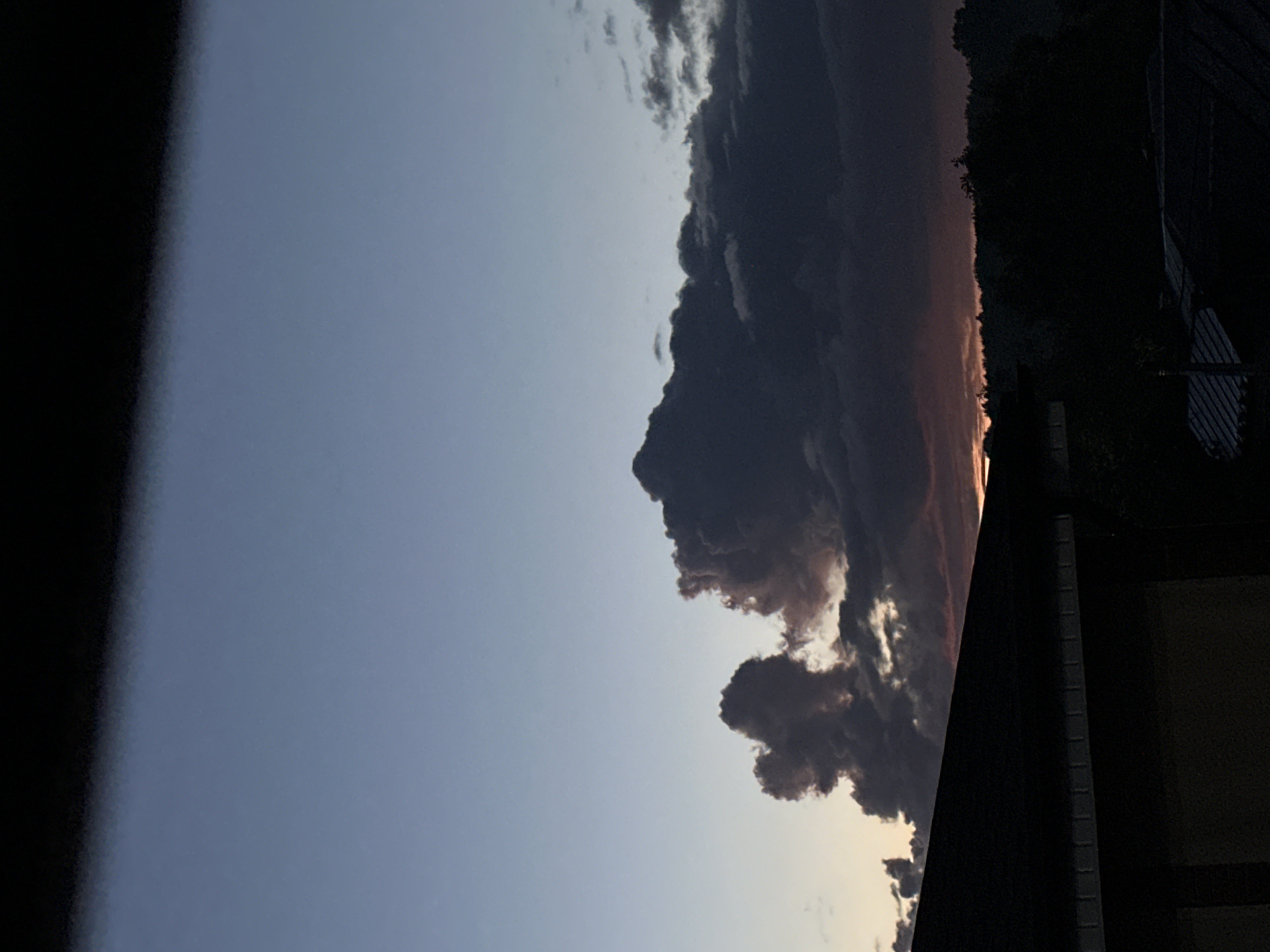 This is a photograph taken in Nizhny Novgorod, Russia, during the twilight hours. The sky is a gradient of colors, transitioning from a pale blue at the top to warmer tones near the horizon. Dark, heavy clouds dominate the middle section of the image, with some lighter clouds scattered throughout. A faint sunset glow can be seen peeking through the clouds near the horizon. In the foreground, the dark silhouette of a roofline is visible, suggesting an urban or suburban setting. The overall atmosphere is calm and serene, typical of a sunset or early evening.