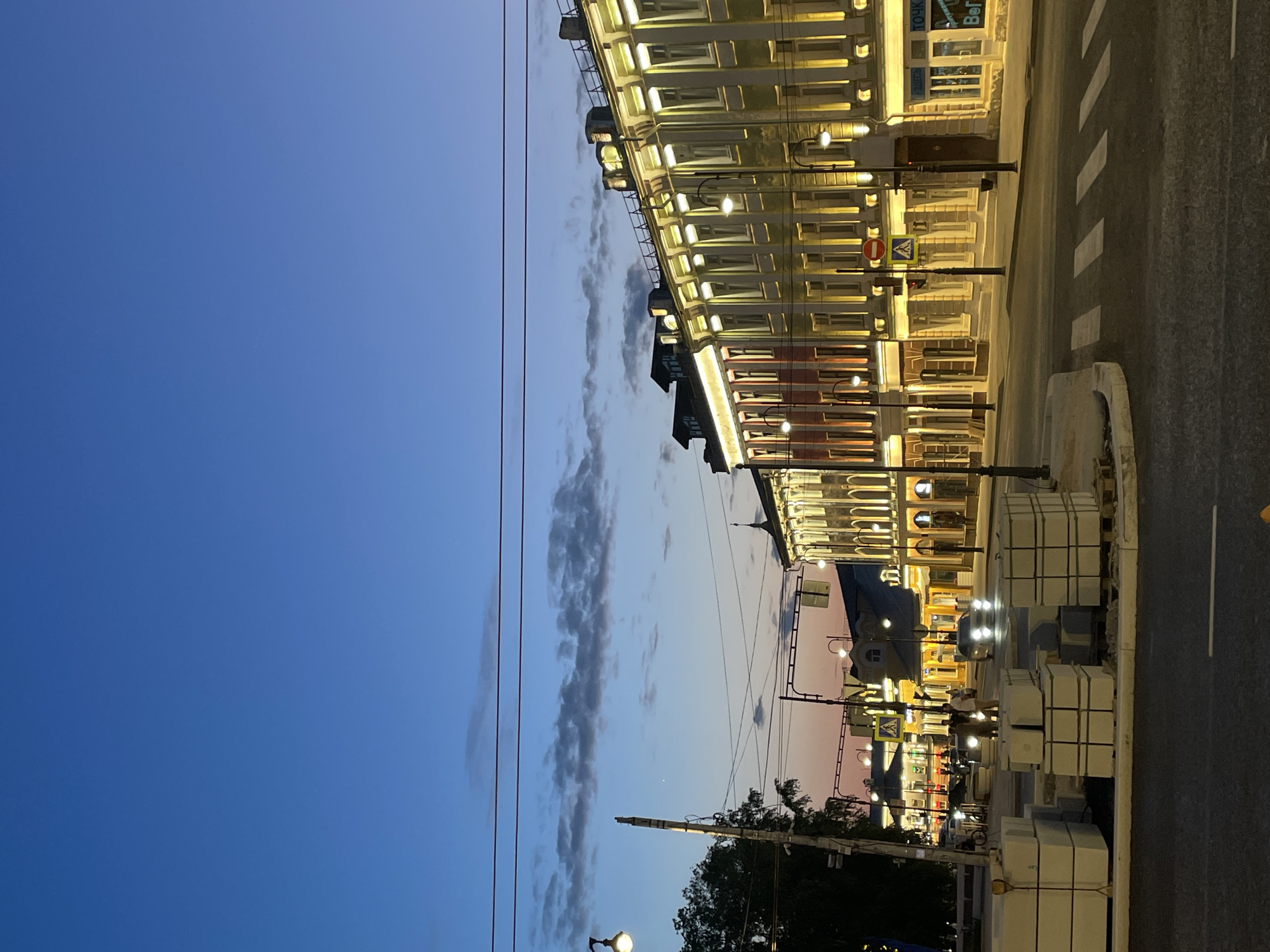 The image depicts a twilight scene on a city street, likely in Vologda, Russia. The sky is a deep blue, transitioning to hues of orange and pink near the horizon, with scattered clouds. On the right side of the frame, a row of ornate buildings lines the street, their facades illuminated by warm yellow lights. The architecture features multiple stories, arched windows, and decorative cornices. Streetlights with glowing globes are visible along the sidewalk.
In the foreground, a street intersection with a pedestrian crossing is visible. Several white concrete blocks, possibly construction materials, are stacked on the side of the road. A few cars are present, with headlights on, suggesting ongoing traffic. A lamppost with wires stretches across the upper portion of the image. In the middle ground, there are pedestrians walking on the sidewalk in front of the illuminated buildings. The overall atmosphere is one of a city transitioning from day to night, with illuminated architecture and visible street life.