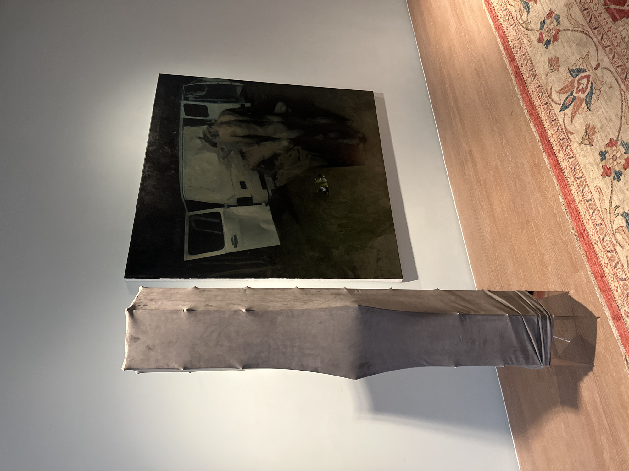 A brightly lit art gallery exhibits two contemporary pieces: a large, dark-toned painting depicting a figure near an opened van, and below it, an elongated, grey, draped form suggestive of a covered object or sculpture. An ornate rug covers the lower right section of the floor. The gallery wall is a light grey. This photograph was likely taken in London, United Kingdom.