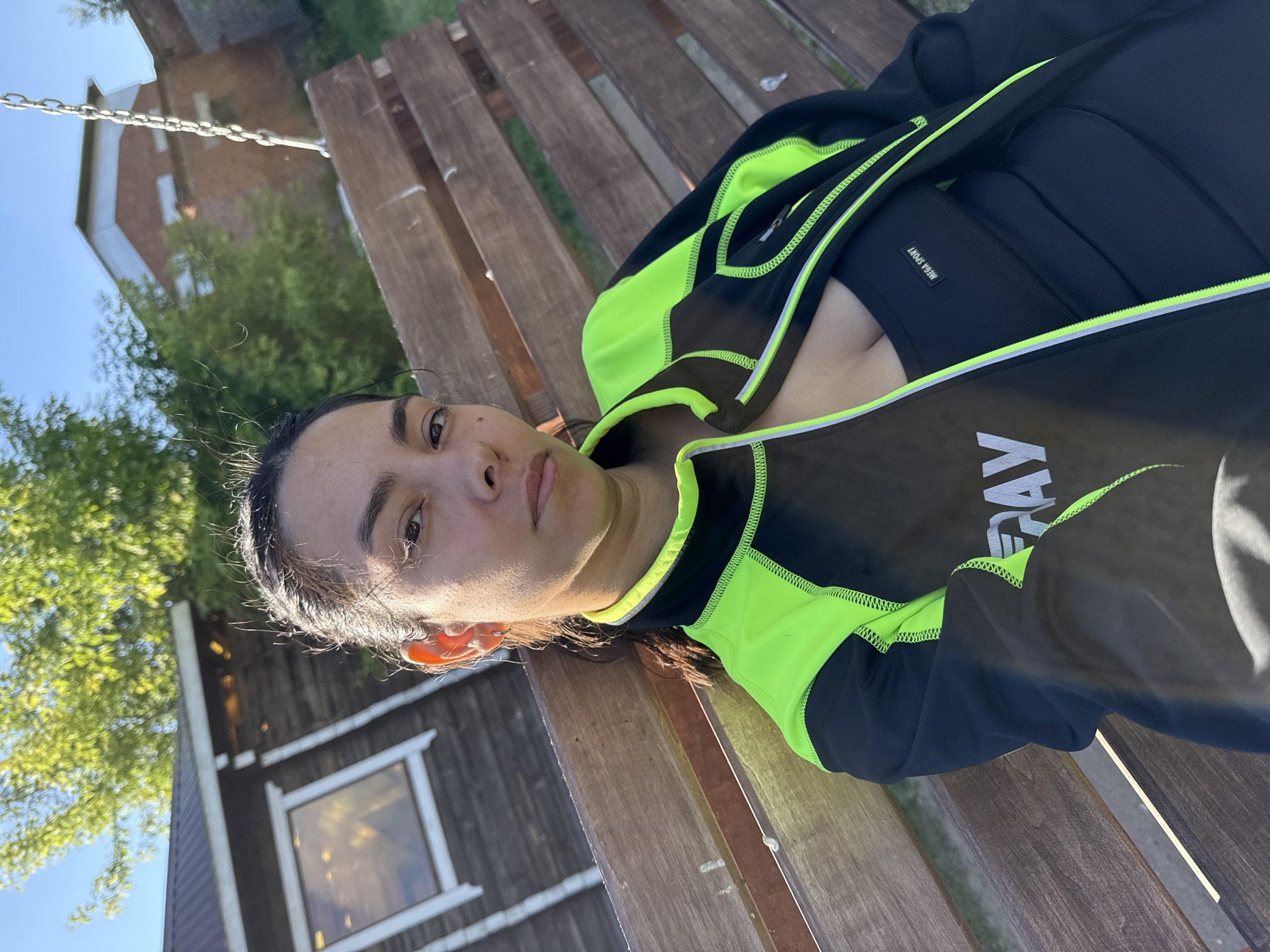 In a daytime outdoor shot, a woman with dark hair pulled back is seated on a wooden swing in what appears to be a backyard or park setting. She is wearing a black and neon green athletic jacket over a black sports bra with the label "MEGA SPORT". A wooden building with a dark roof and a window is visible to her left. In the background, a brick building and trees are present under a clear blue sky. The scene is located in the city of Chaikovsky, Russia.
