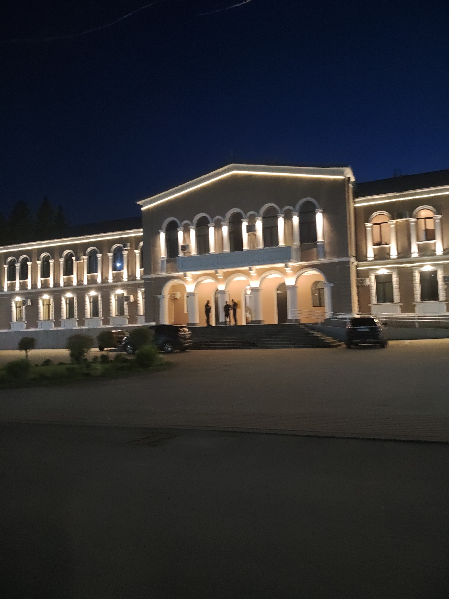 A wide shot, taken at night, shows the front of a large, illuminated building. The building is light-colored with arched windows and decorative lighting along the roofline. In front of the building, there is a paved area with several cars parked. A few people are visible on the steps leading up to the building's entrance. The sky above is a deep blue, suggesting it is either early evening or late night. The overall scene appears to be a public building or institution in the city of Terjola, Georgia.
